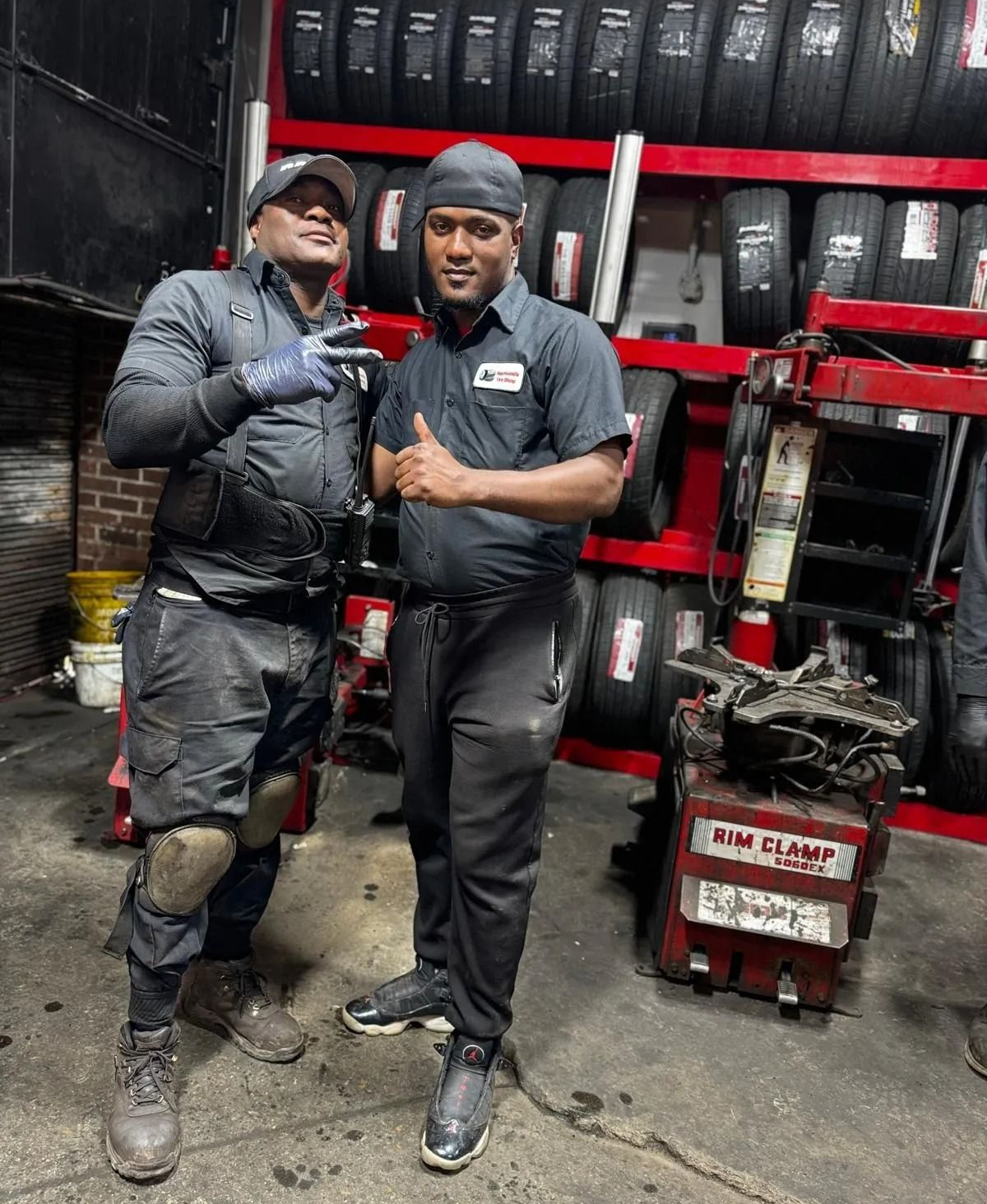Two auto mechanics, one with a peace sign, in a tire shop. Tires line the wall.