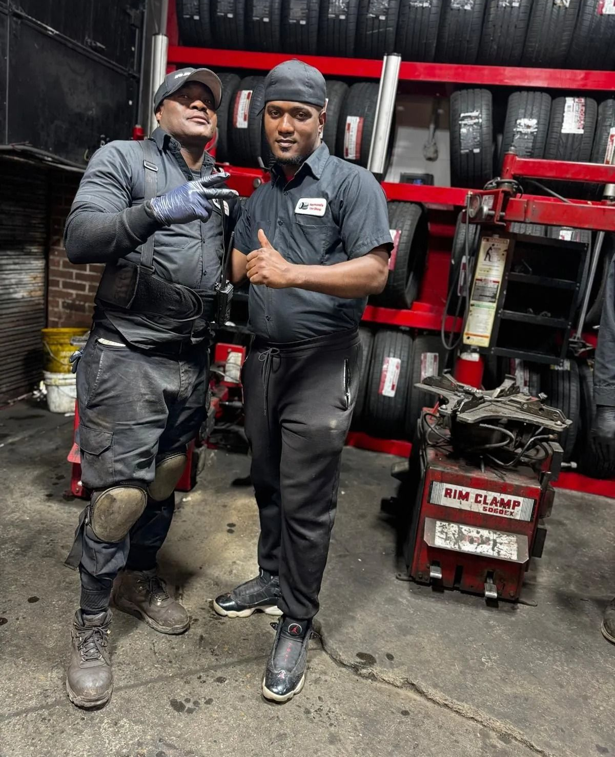 Two auto mechanics, one with a peace sign, in a tire shop. Tires line the wall.