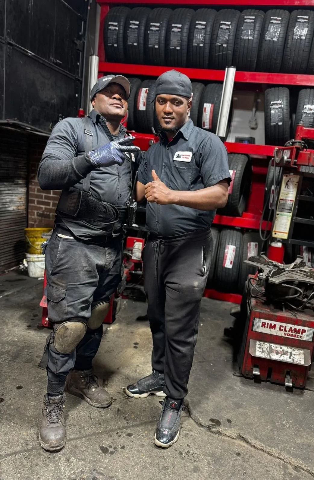 Two mechanics in a tire shop pose; one gives a peace sign, the other a thumbs up. Tires are in the background.