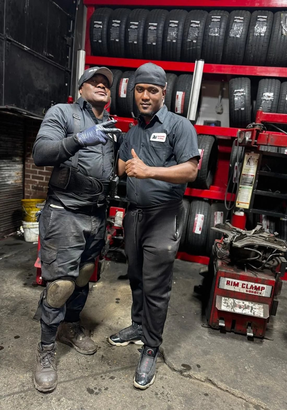 Two mechanics in a tire shop pose; one gives a peace sign, the other a thumbs up. Tires are in the background.