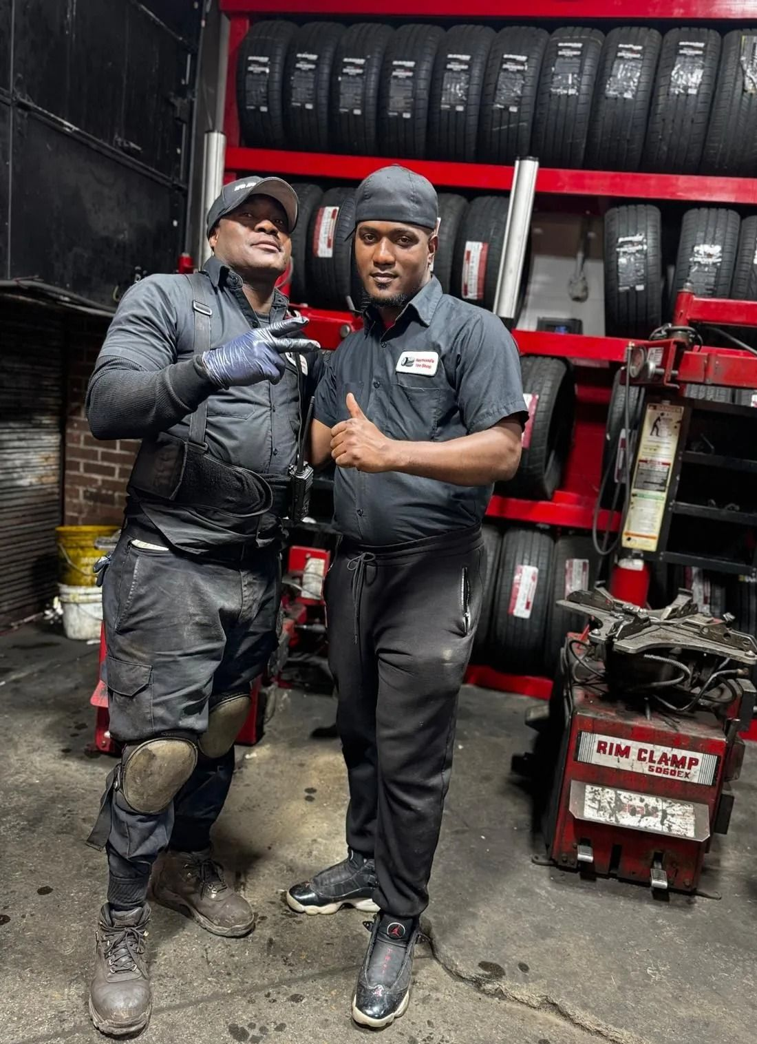 Two mechanics in a tire shop pose; one gives a peace sign, the other a thumbs up. Tires are in the background.
