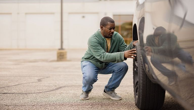Person crouches beside a parked car, inspecting or working near the front wheel in a lot.
