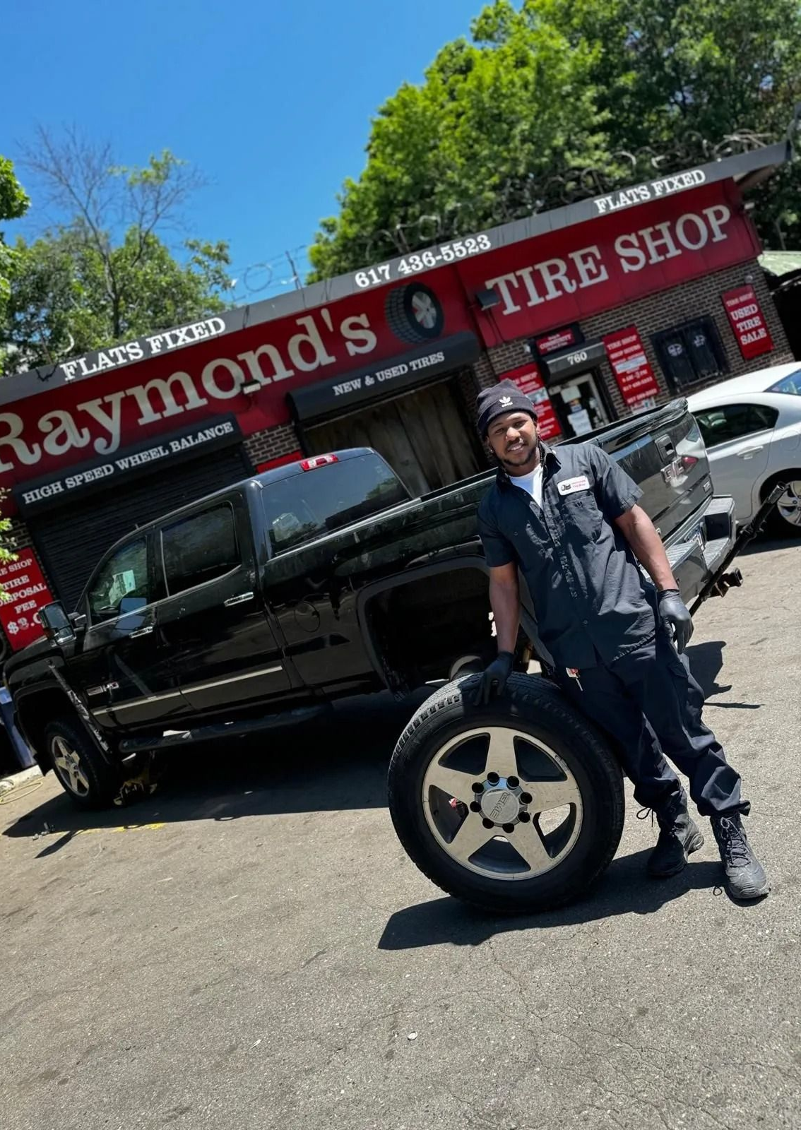 Mechanic poses with a tire in front of Raymond's Tire Shop with a black pickup truck.