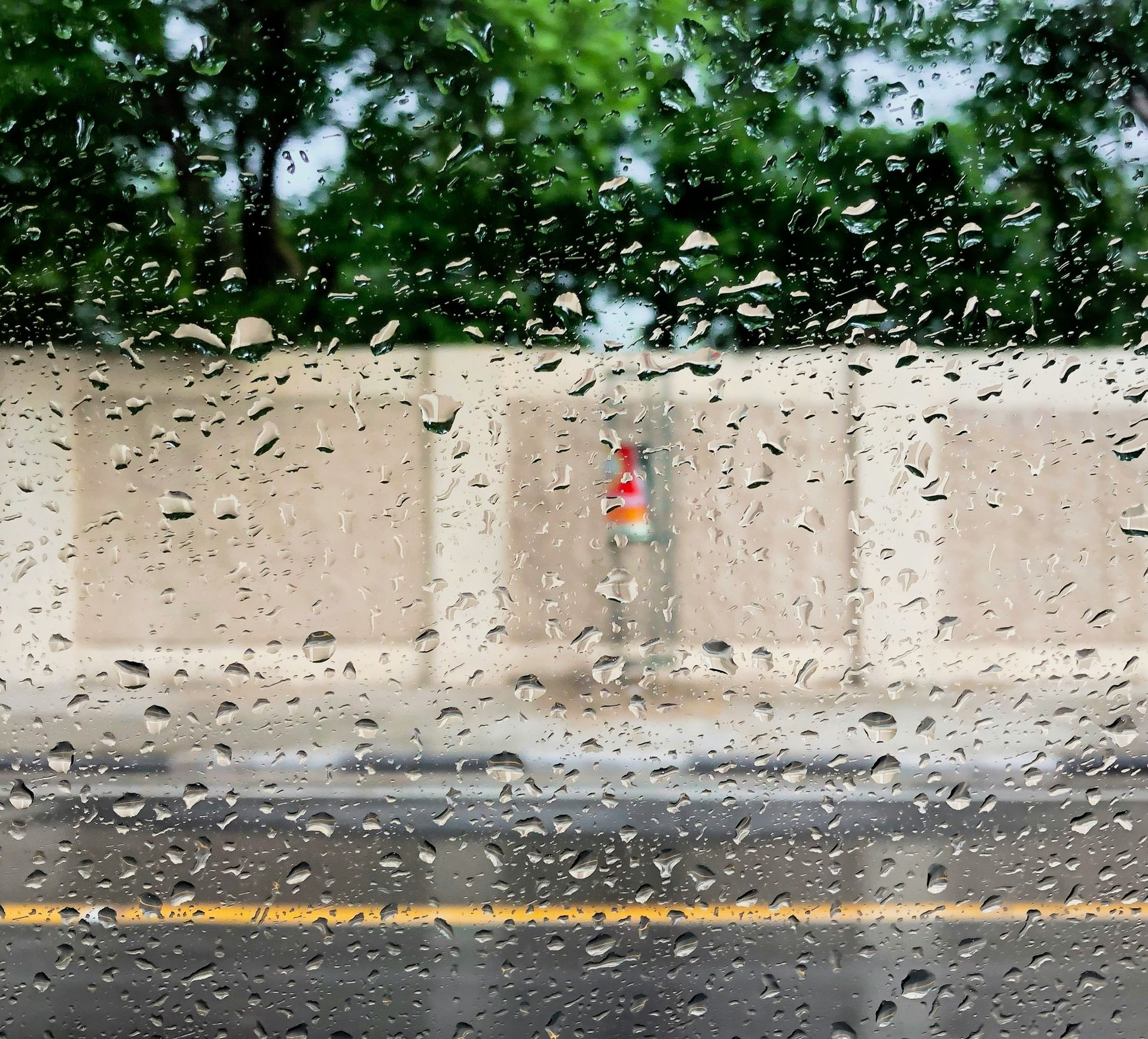 Rain-soaked street seen through a window with water droplets, beige wall, and blurred trees beyond.