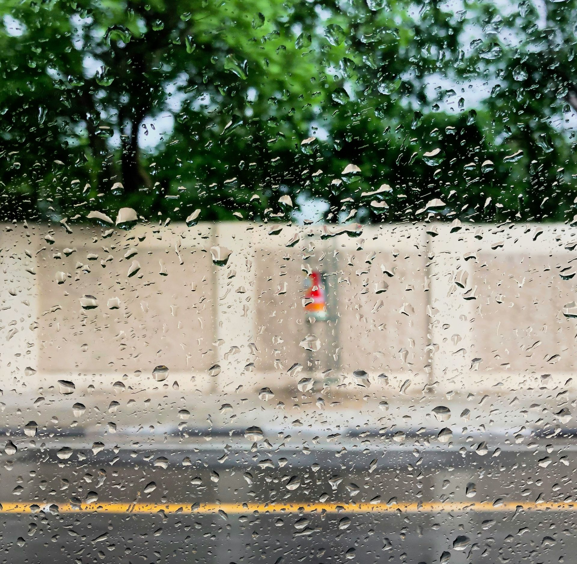 Rain-soaked street seen through a window with water droplets, beige wall, and blurred trees beyond.