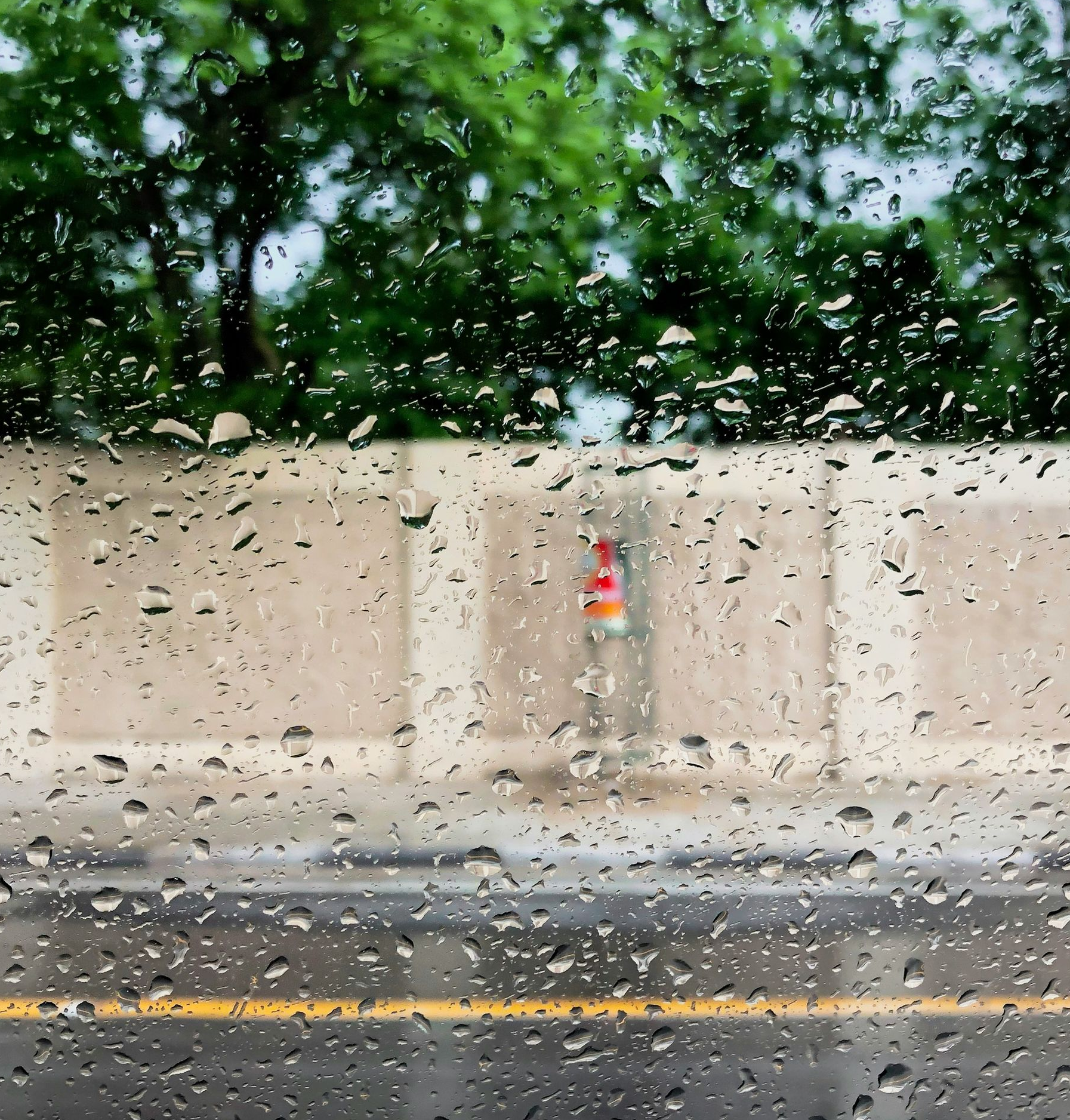 Rain-soaked street seen through a window with water droplets, beige wall, and blurred trees beyond.