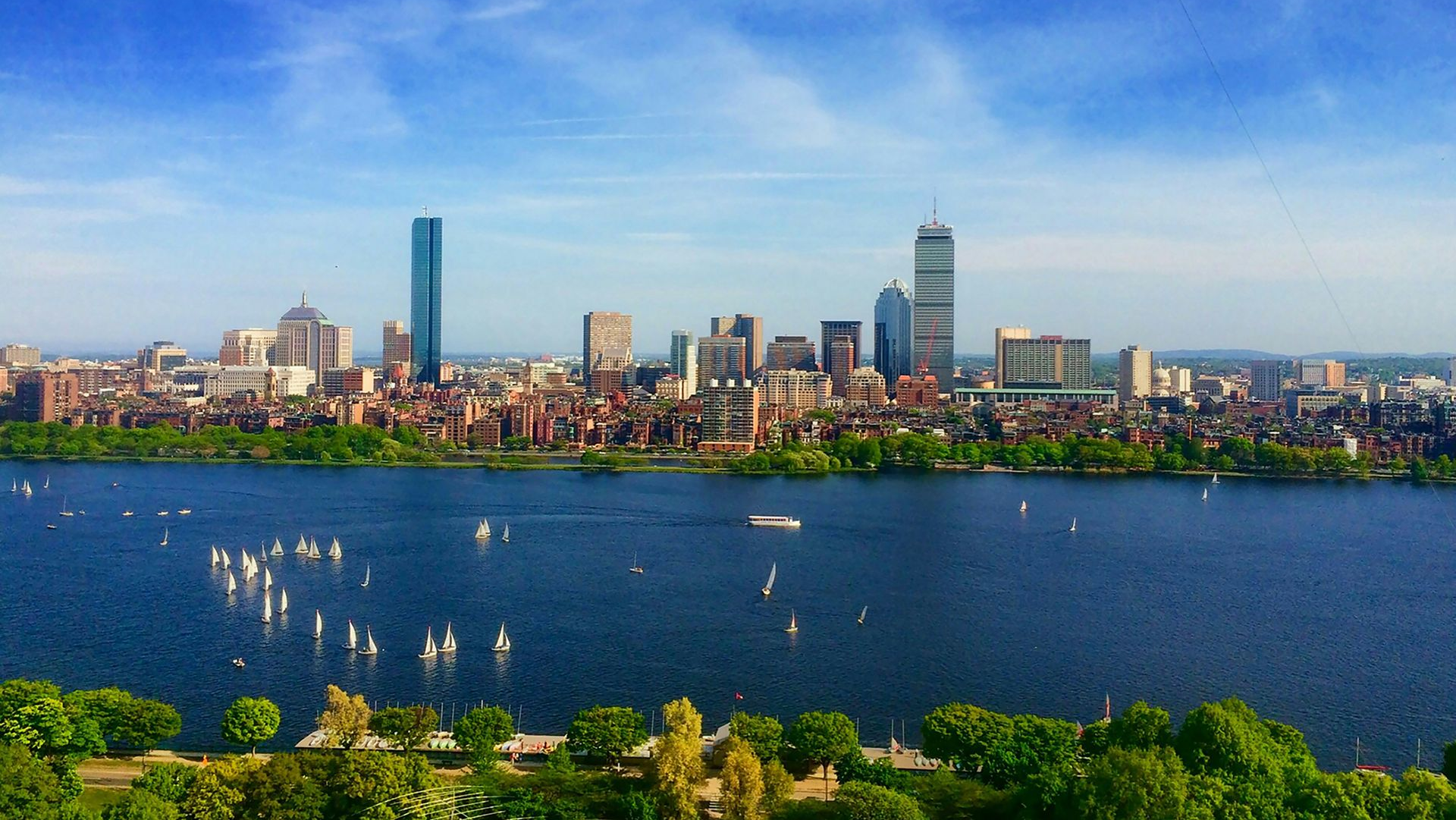 A panoramic view of the Boston skyline across the Charles River, featuring sailboats on the water under a clear blue sky.