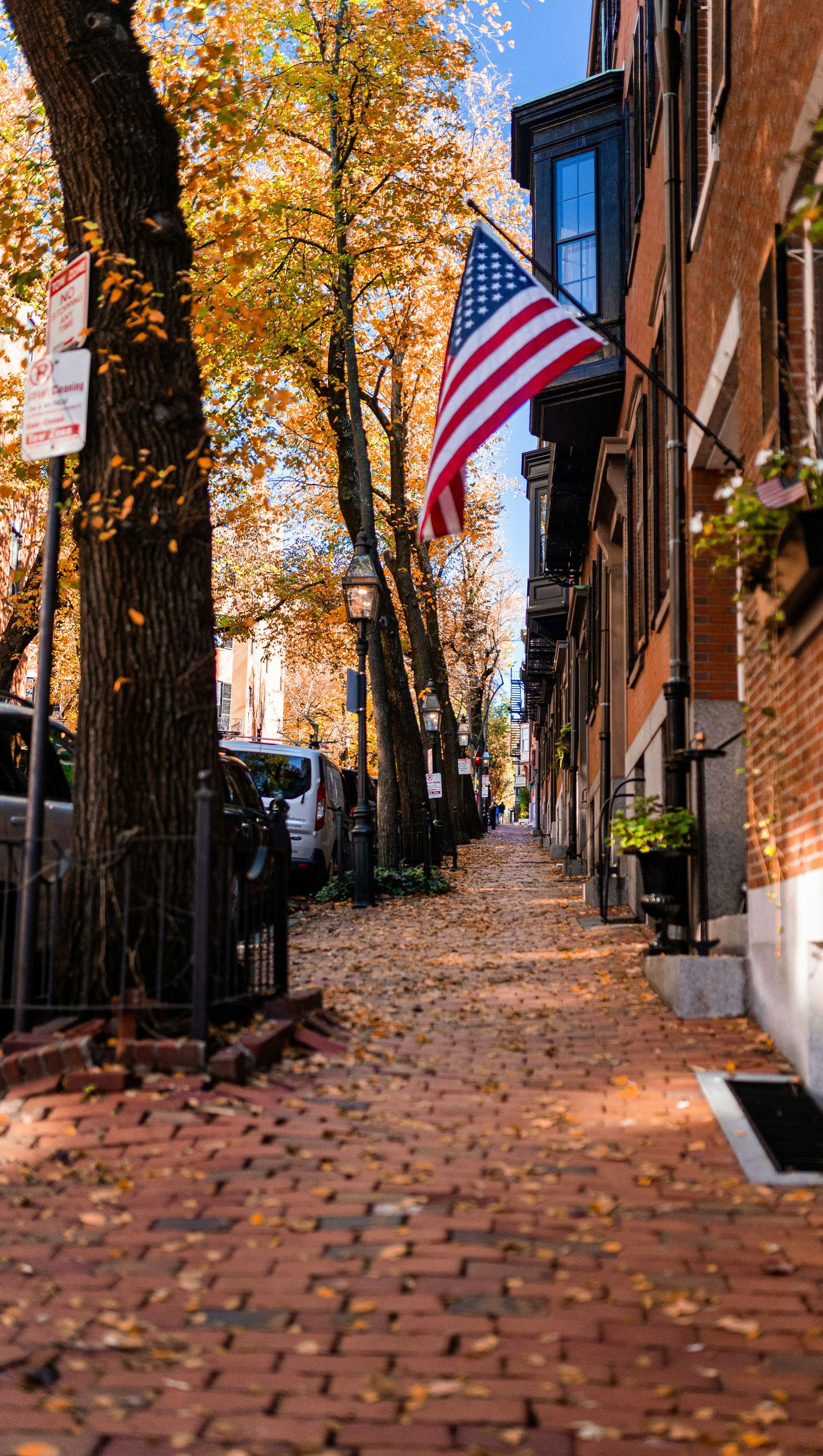 A brick sidewalk lined with autumn trees and a brick building displaying an American flag on a sunny day.