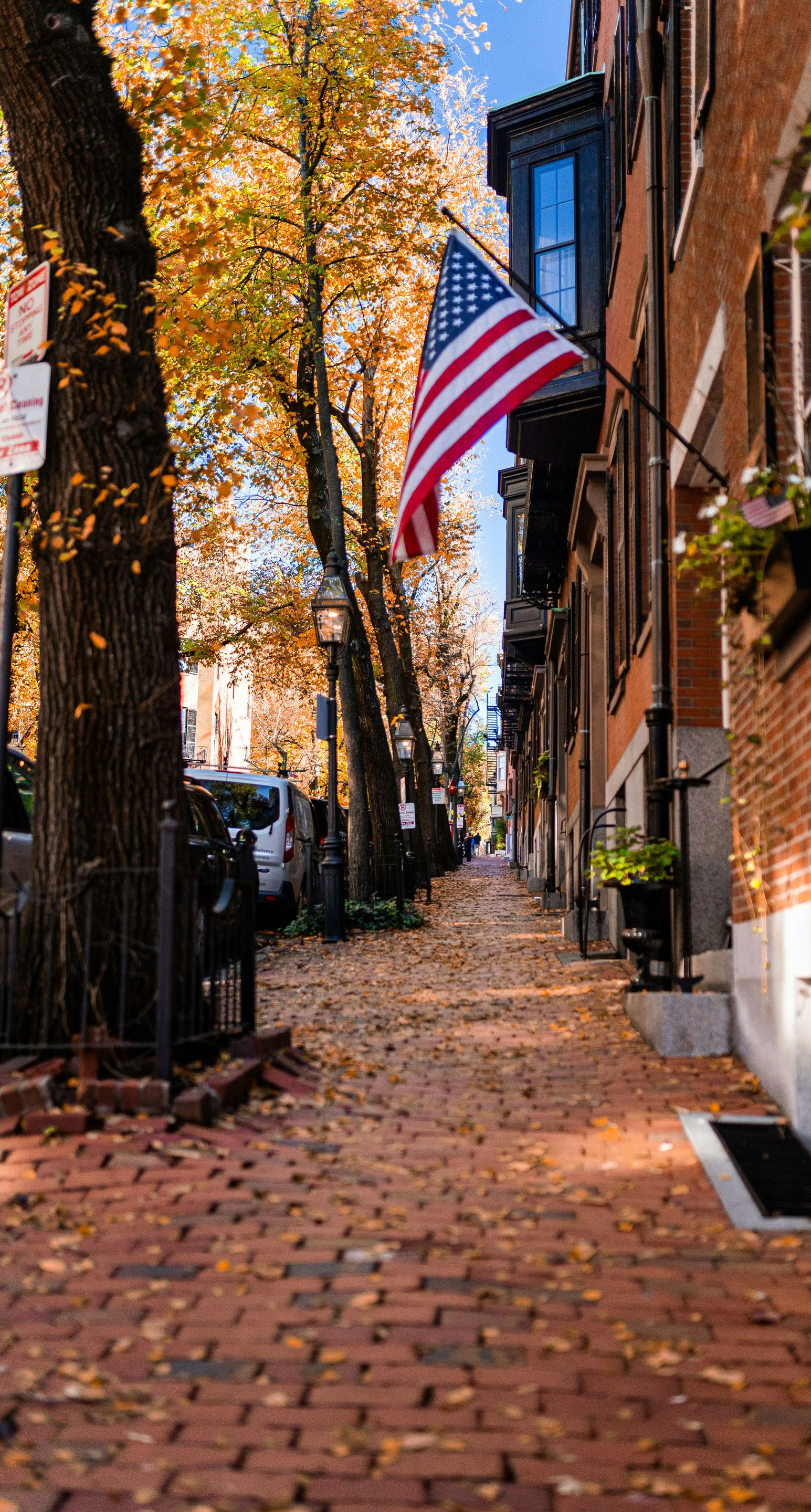 A brick sidewalk lined with autumn trees and a brick building displaying an American flag on a sunny day.