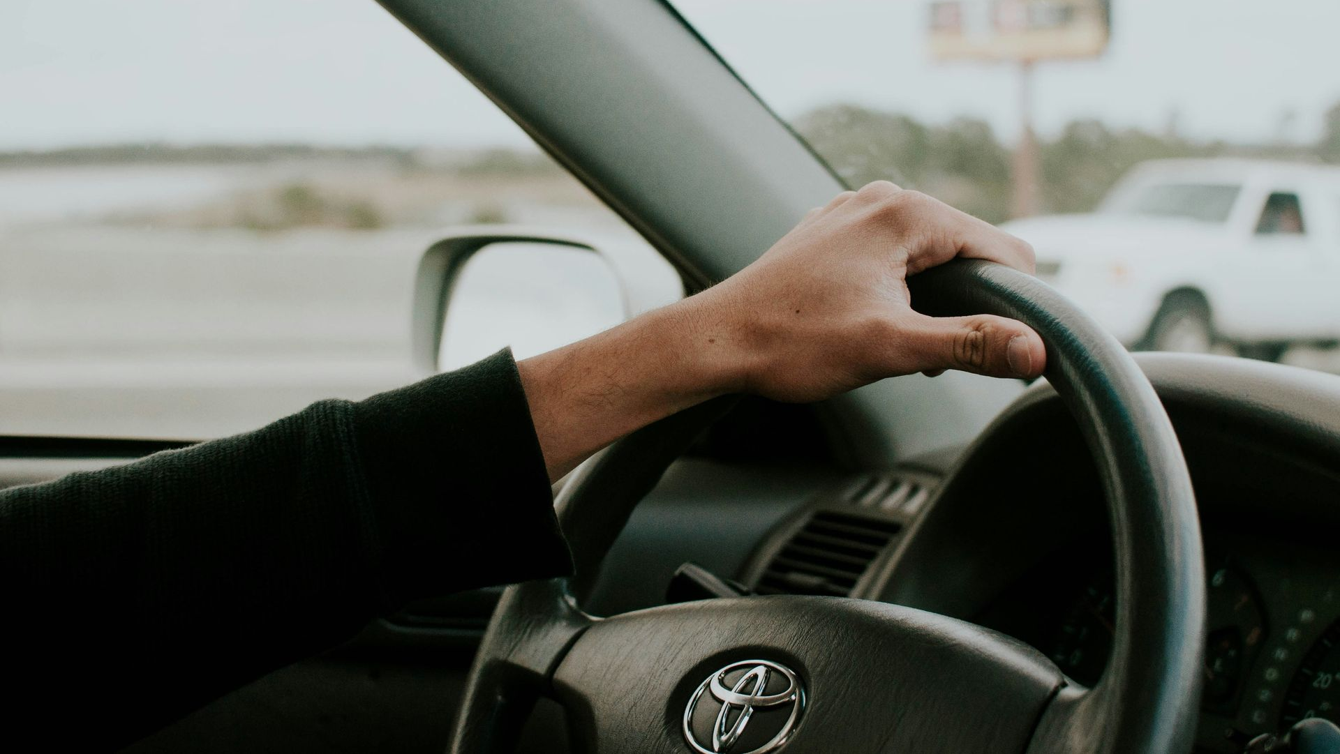 Driver’s hand on steering wheel inside a Toyota car on a road, with another vehicle visible outside.