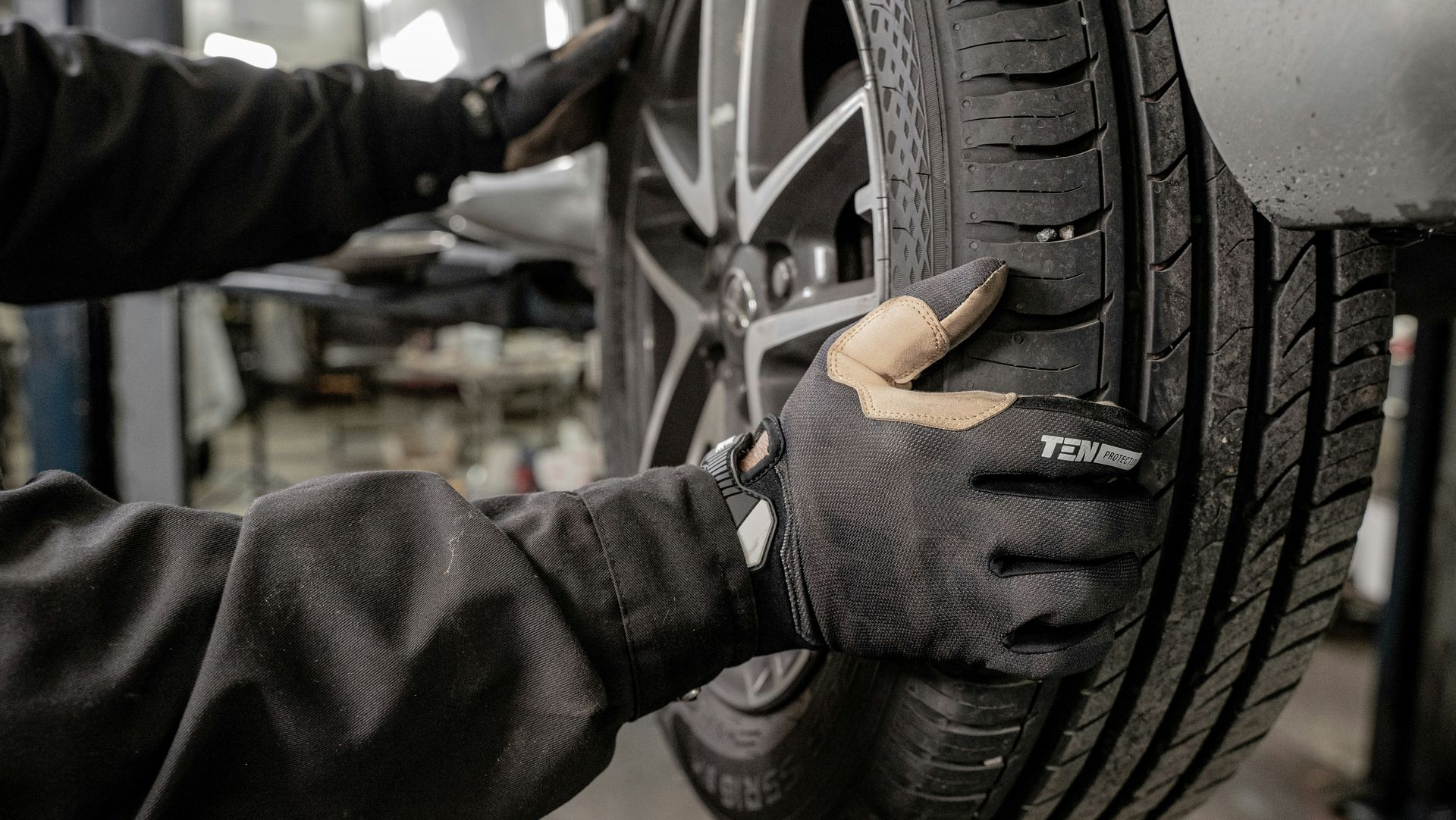 A mechanic in black workwear and gloves inspecting a car tire at a repair shop.