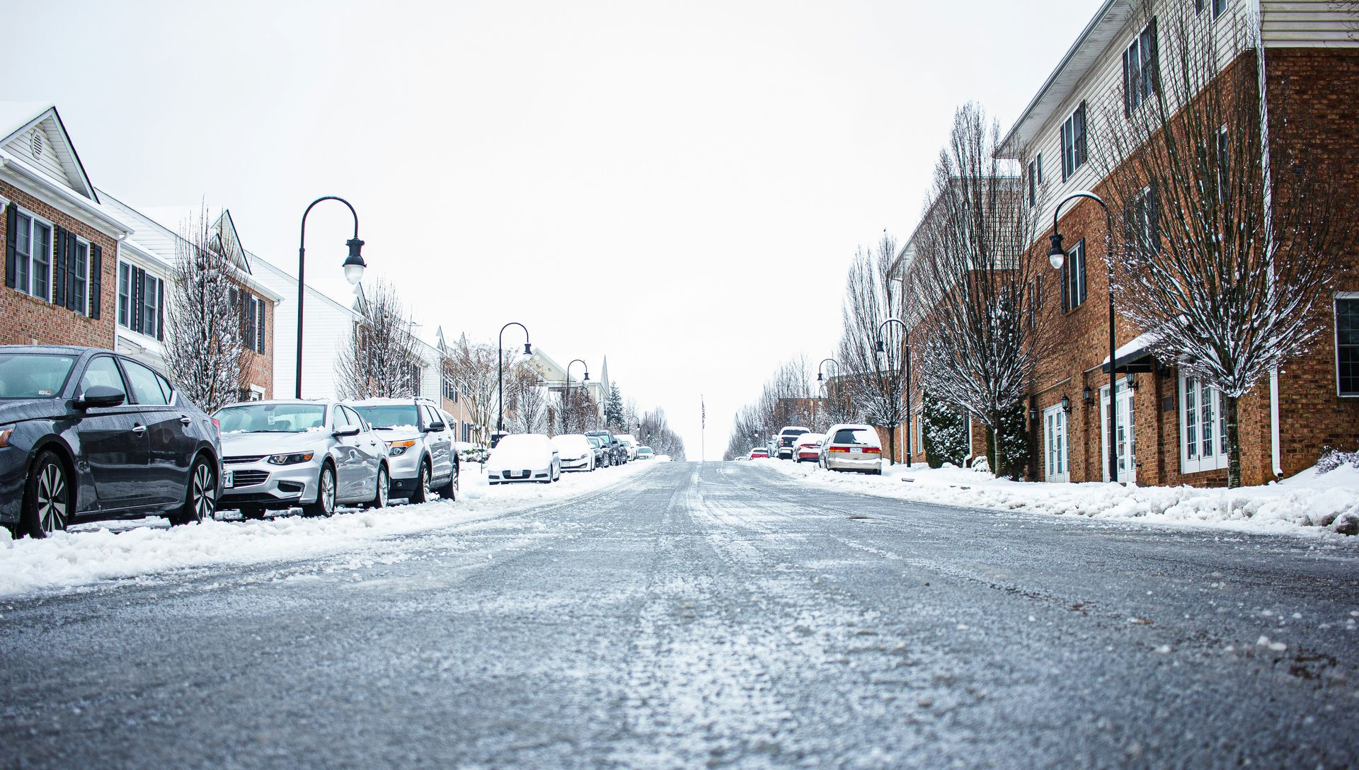 Snowy street with parked cars and buildings, under overcast sky.
