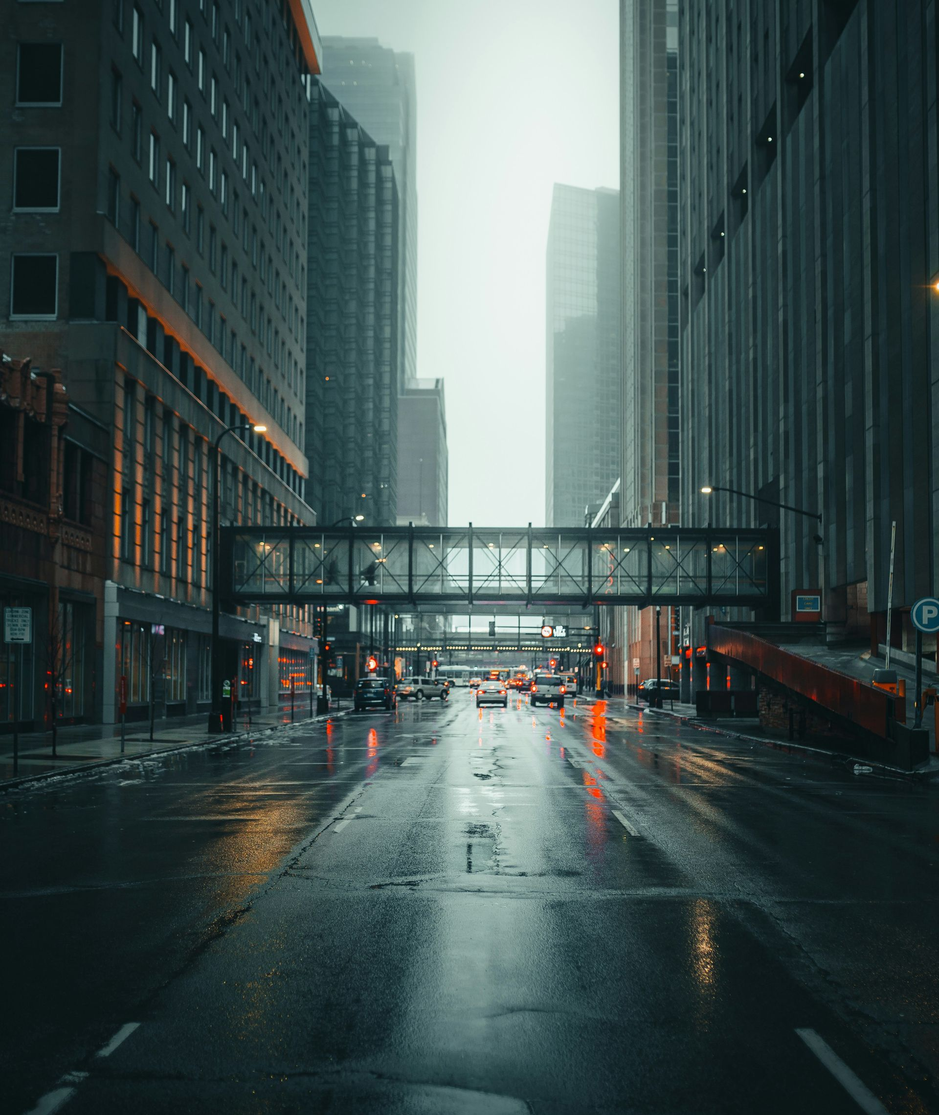 Rainy city street between tall skyscrapers with wet pavement and a pedestrian bridge overhead