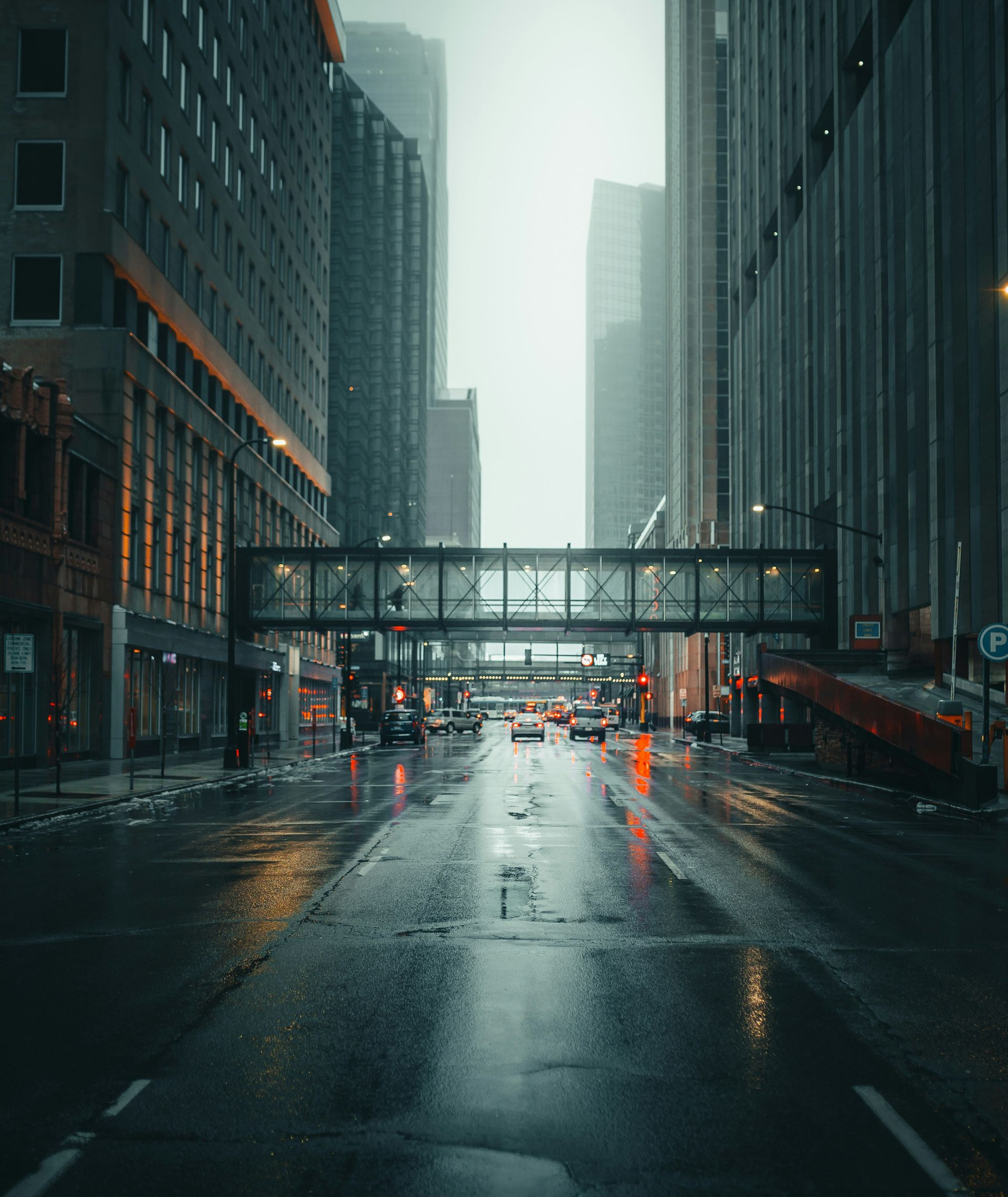 Rainy city street between tall skyscrapers with wet pavement and a pedestrian bridge overhead