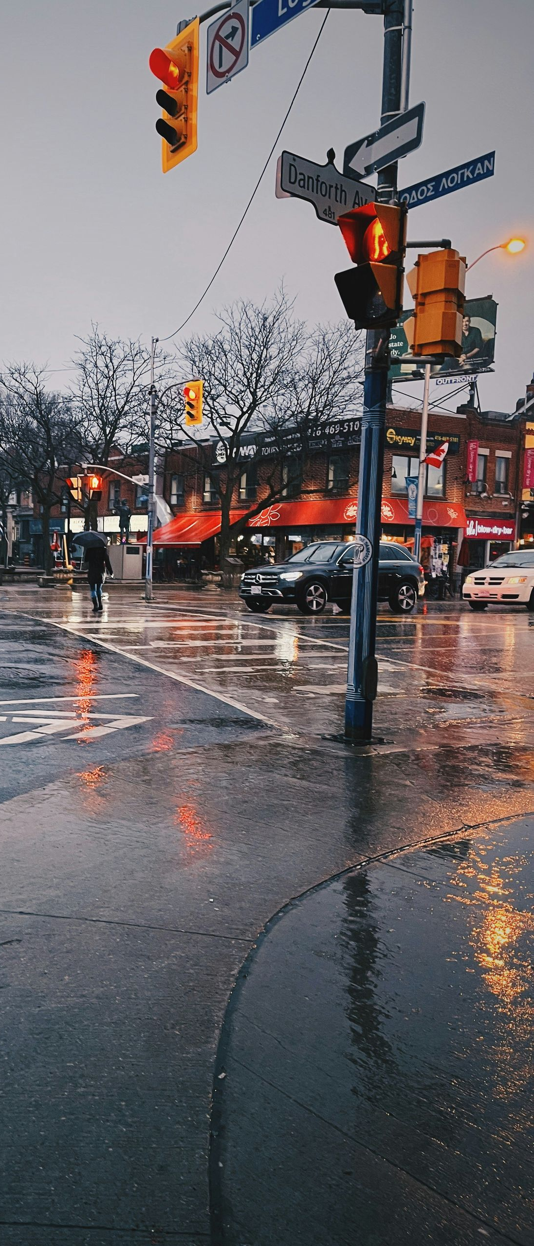 Rainy city street with wet pavement, traffic lights, and storefronts reflecting orange lights.