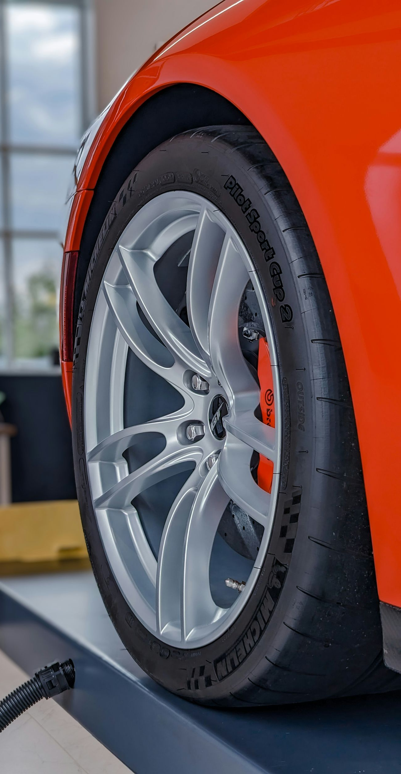 A bright orange car on a mechanic’s lift, showcasing a silver alloy wheel and high-performance tire in a shop.