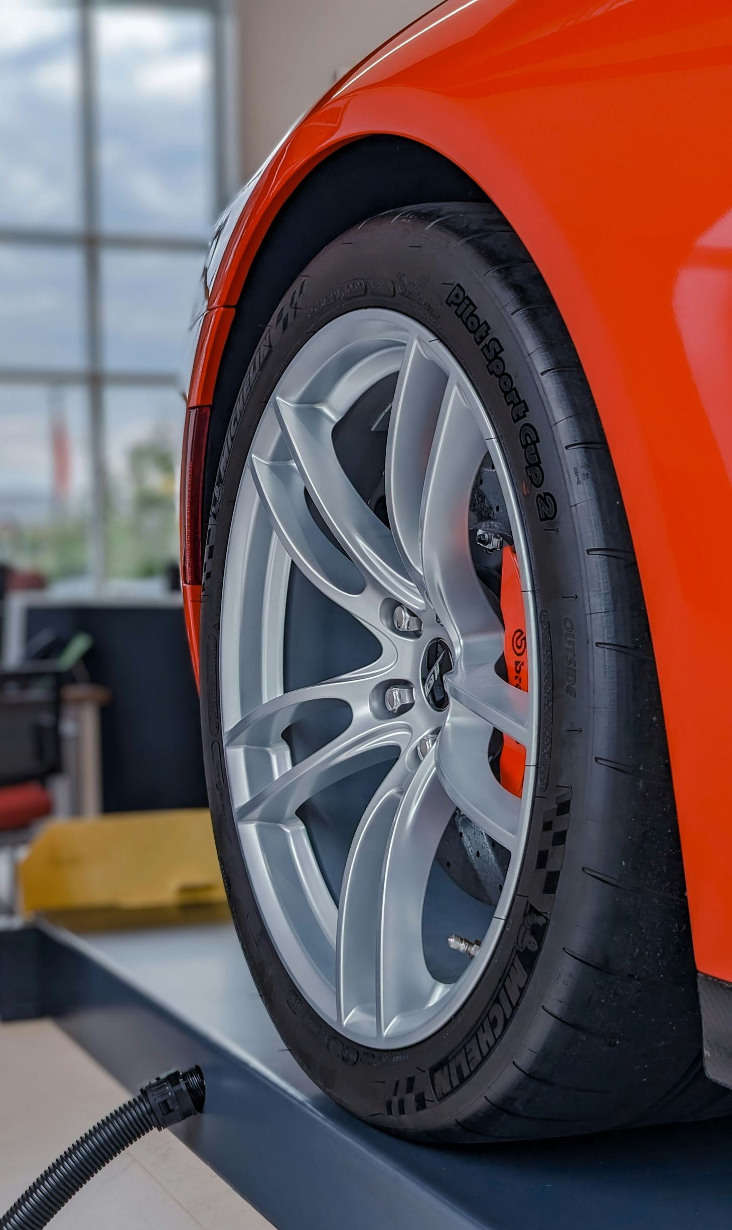 A bright orange car on a mechanic’s lift, showcasing a silver alloy wheel and high-performance tire in a shop.