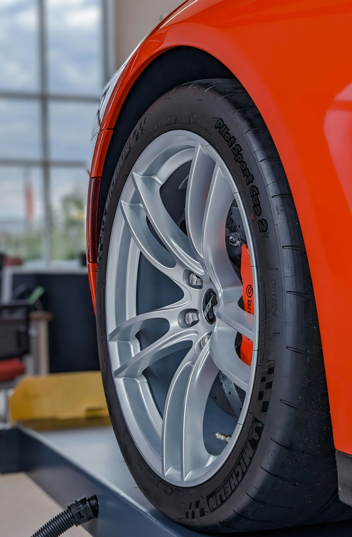 A bright orange car on a mechanic’s lift, showcasing a silver alloy wheel and high-performance tire in a shop.