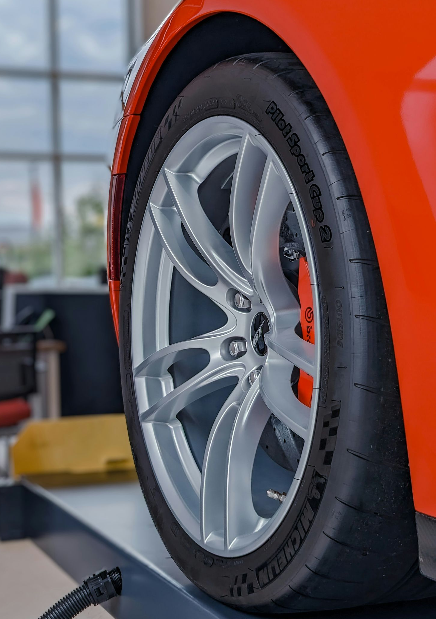 A bright orange car on a mechanic’s lift, showcasing a silver alloy wheel and high-performance tire in a shop.