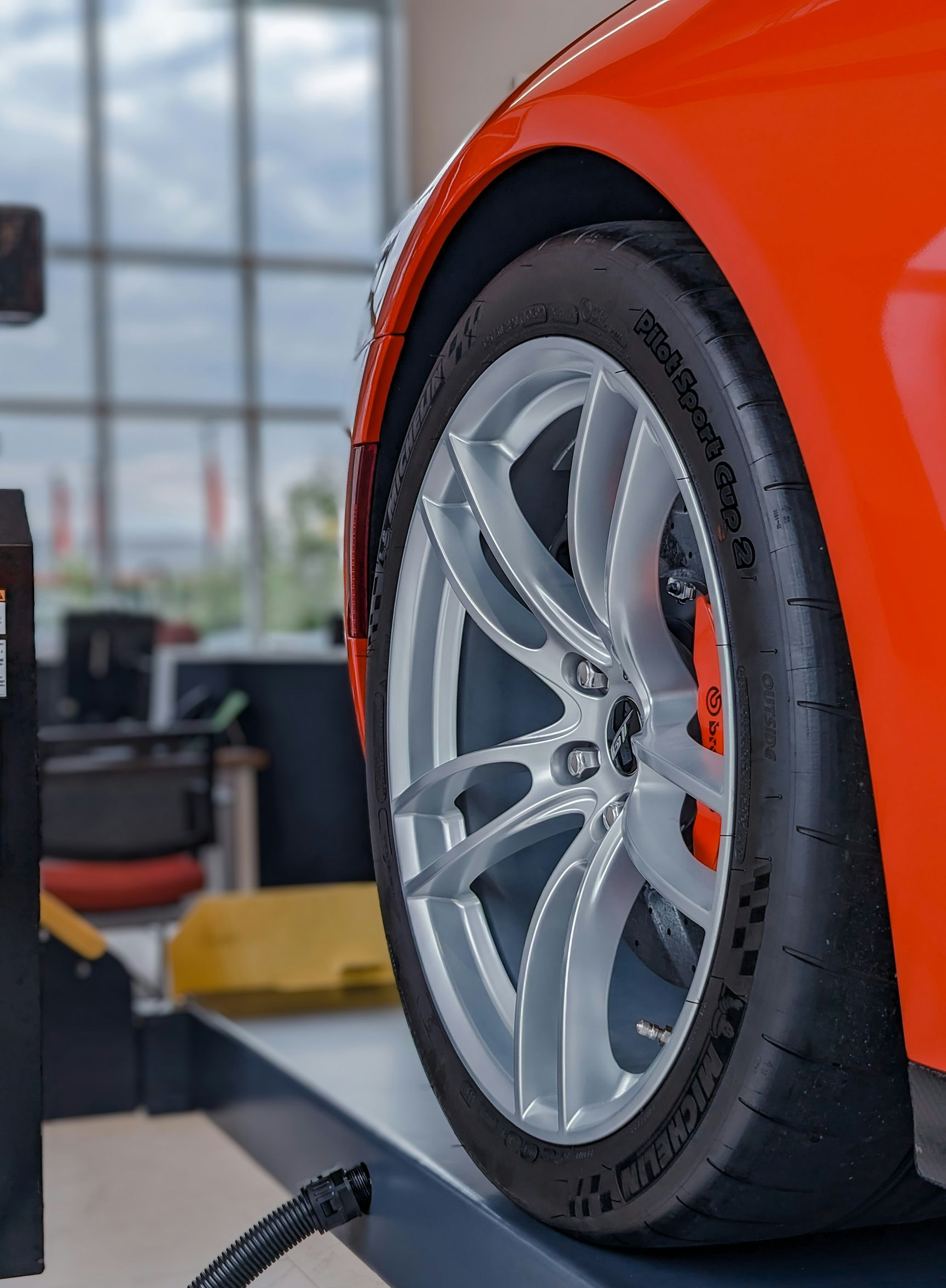A bright orange car on a mechanic’s lift, showcasing a silver alloy wheel and high-performance tire in a shop.