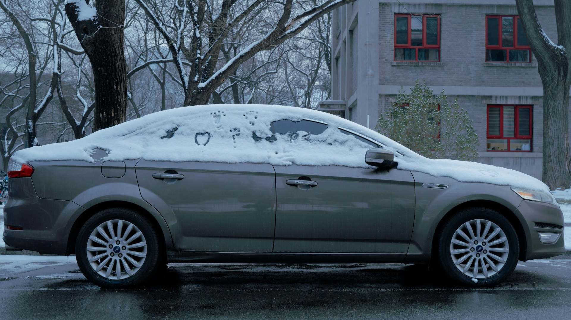 Gray car covered in snow parked on a street in front of a building with red window frames.