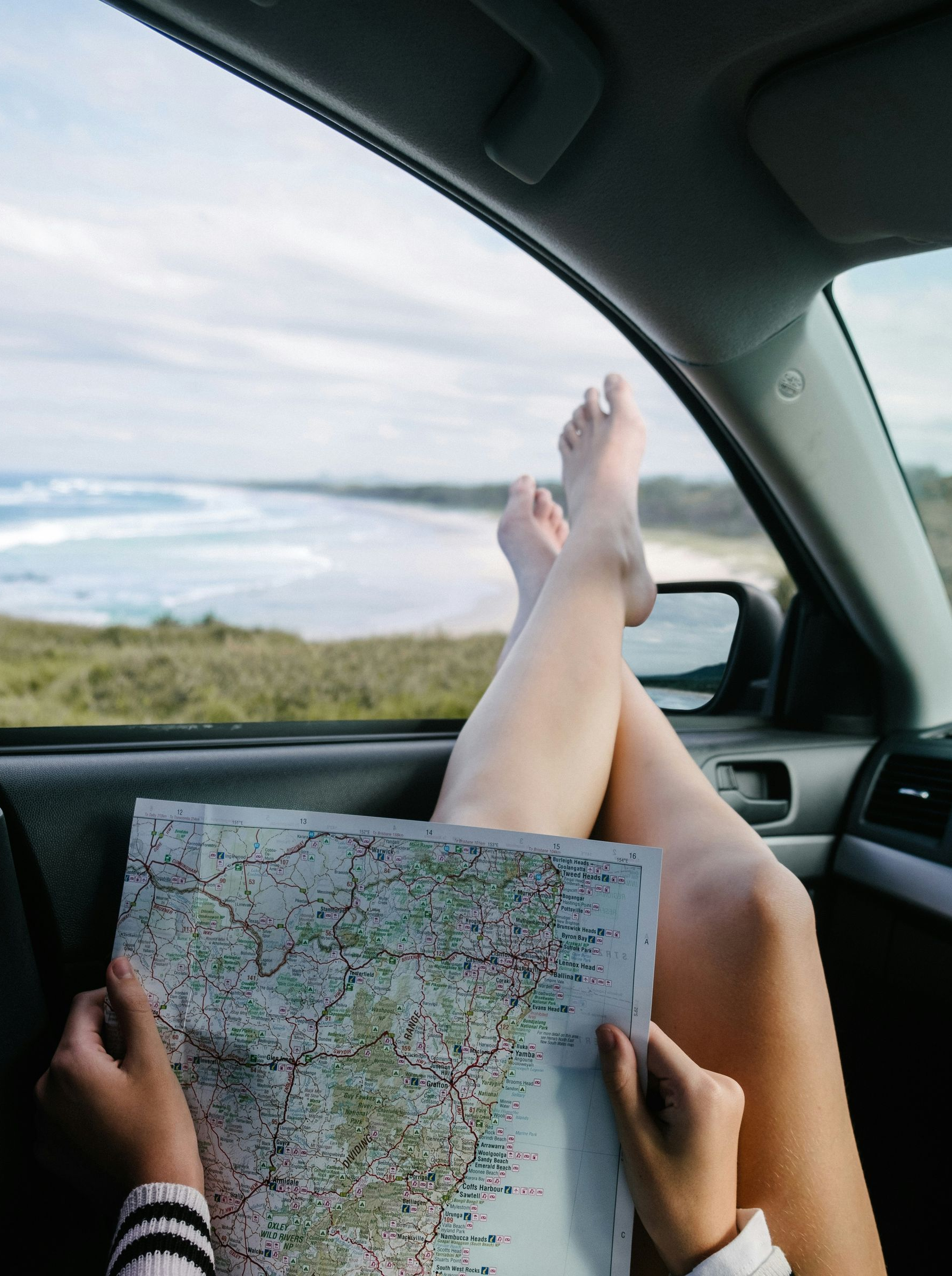 Passenger in a car holds a map while feet rest out the window on a scenic road.