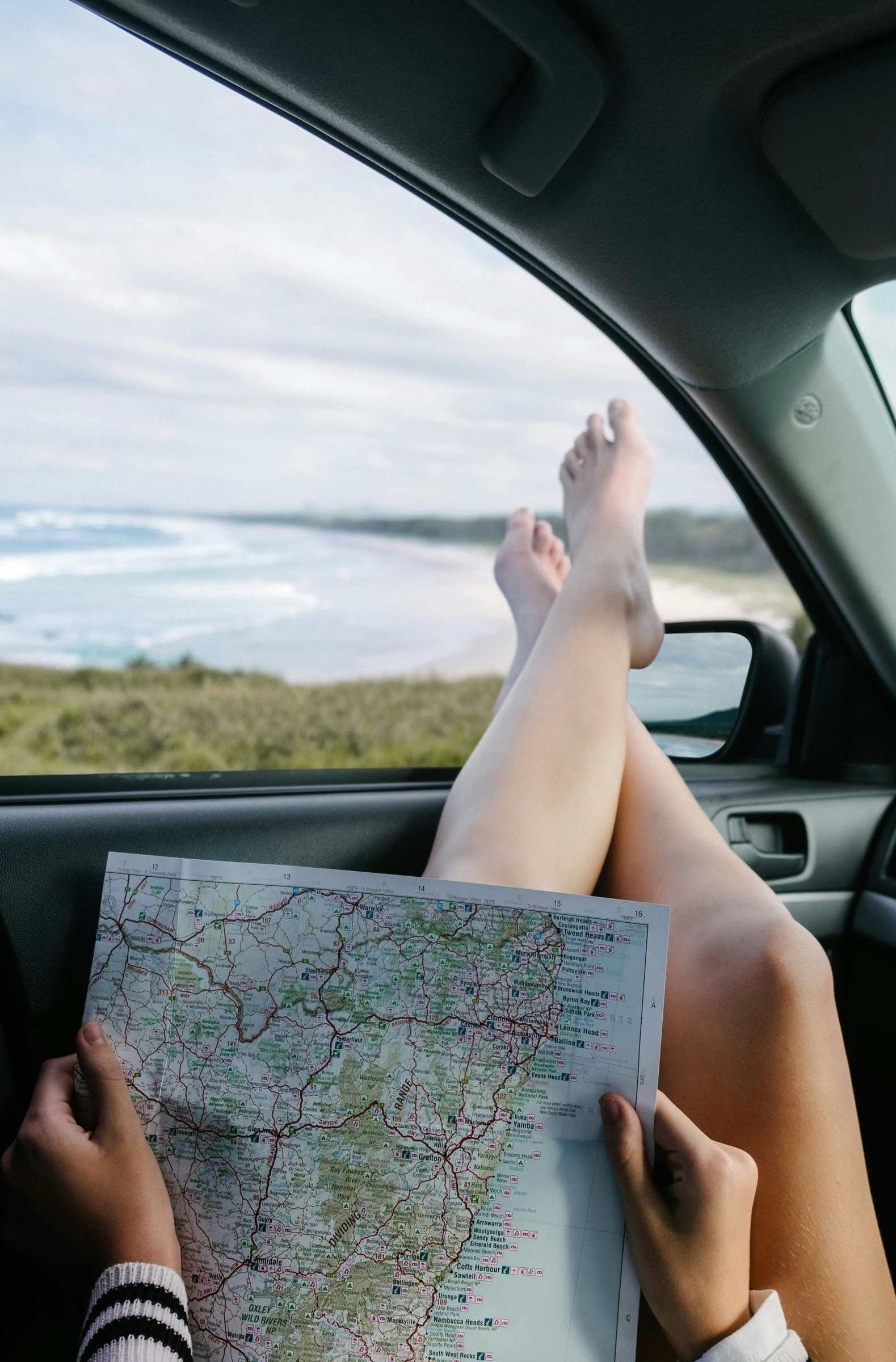 Passenger in a car holds a map while feet rest out the window on a scenic road.