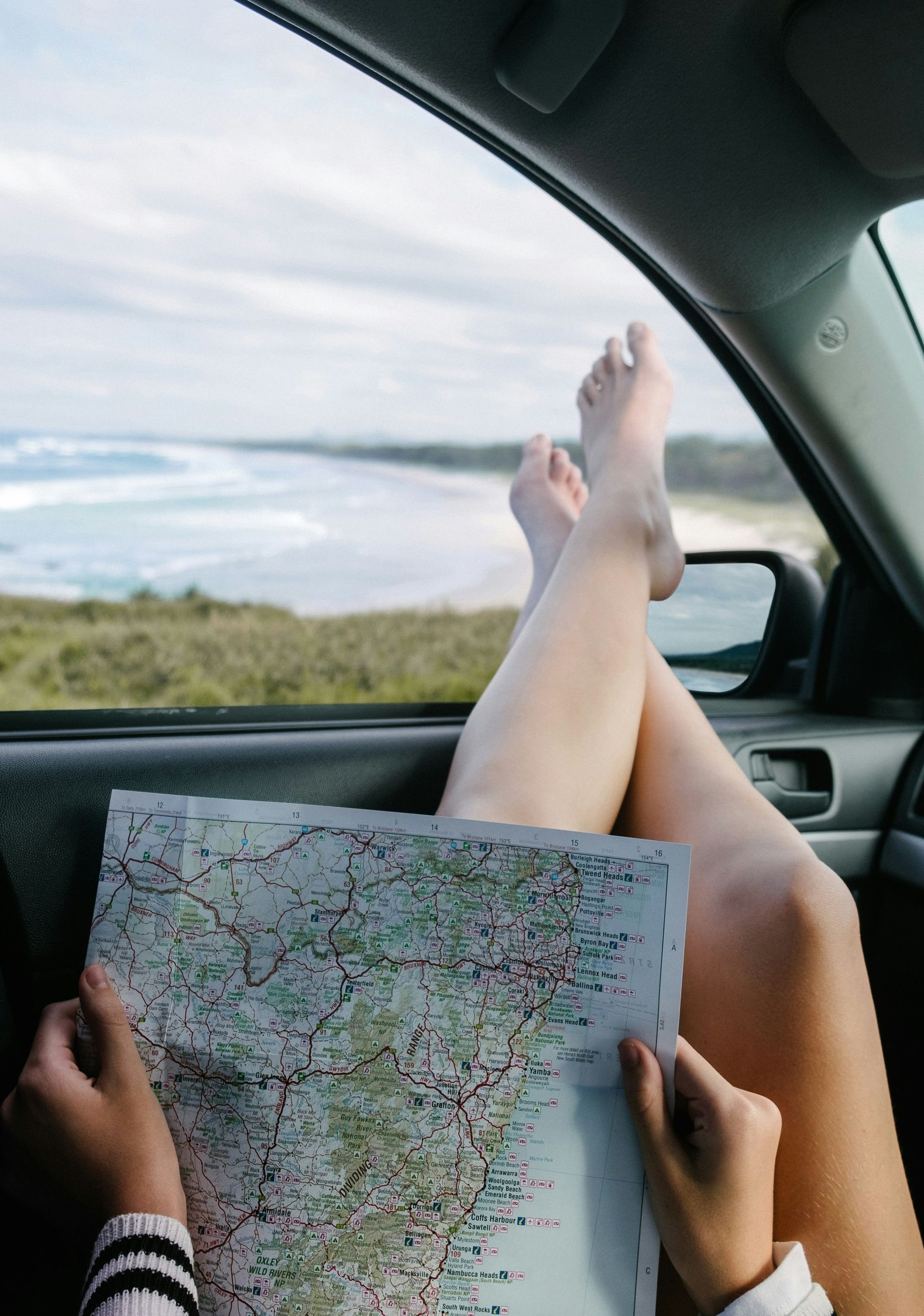 Passenger in a car holds a map while feet rest out the window on a scenic road.