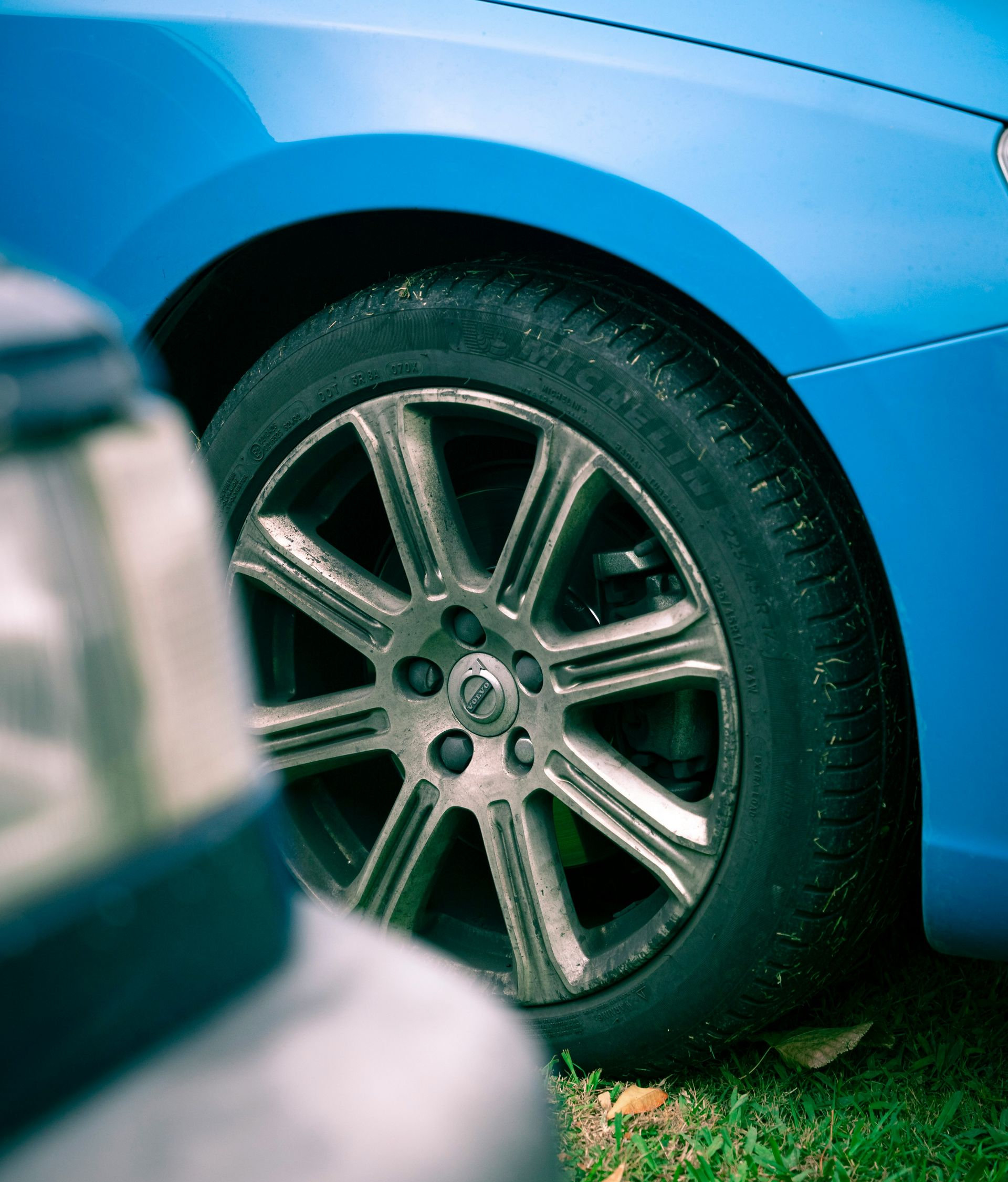 A bright blue car with a multi-spoke metallic wheel parked on grass, partially obscured by a vehicle in the foreground.