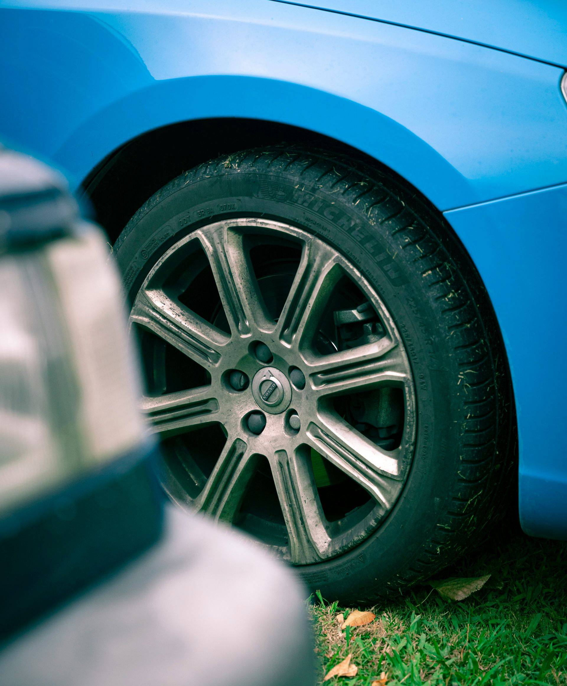 A bright blue car with a multi-spoke metallic wheel parked on grass, partially obscured by a vehicle in the foreground.