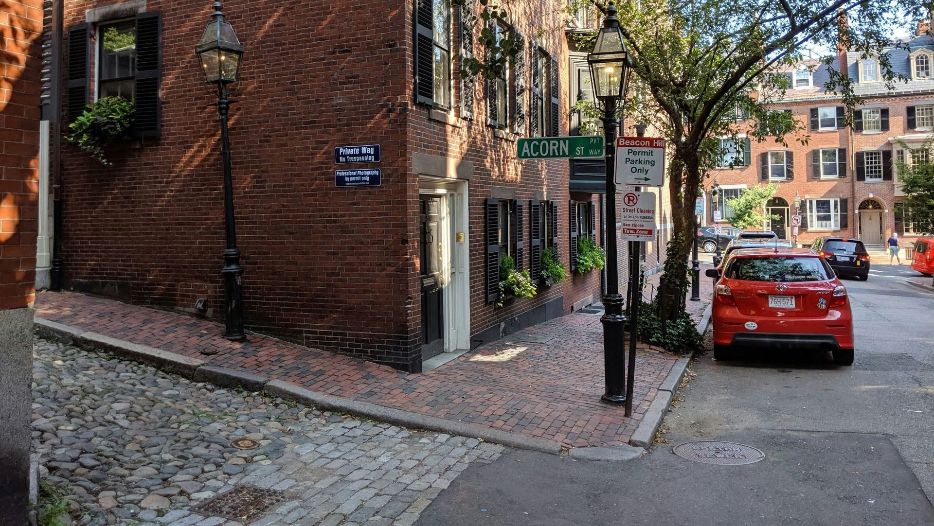A red brick building on a corner in a historic neighborhood, featuring cobblestone paths, street lamps, and a parked car.