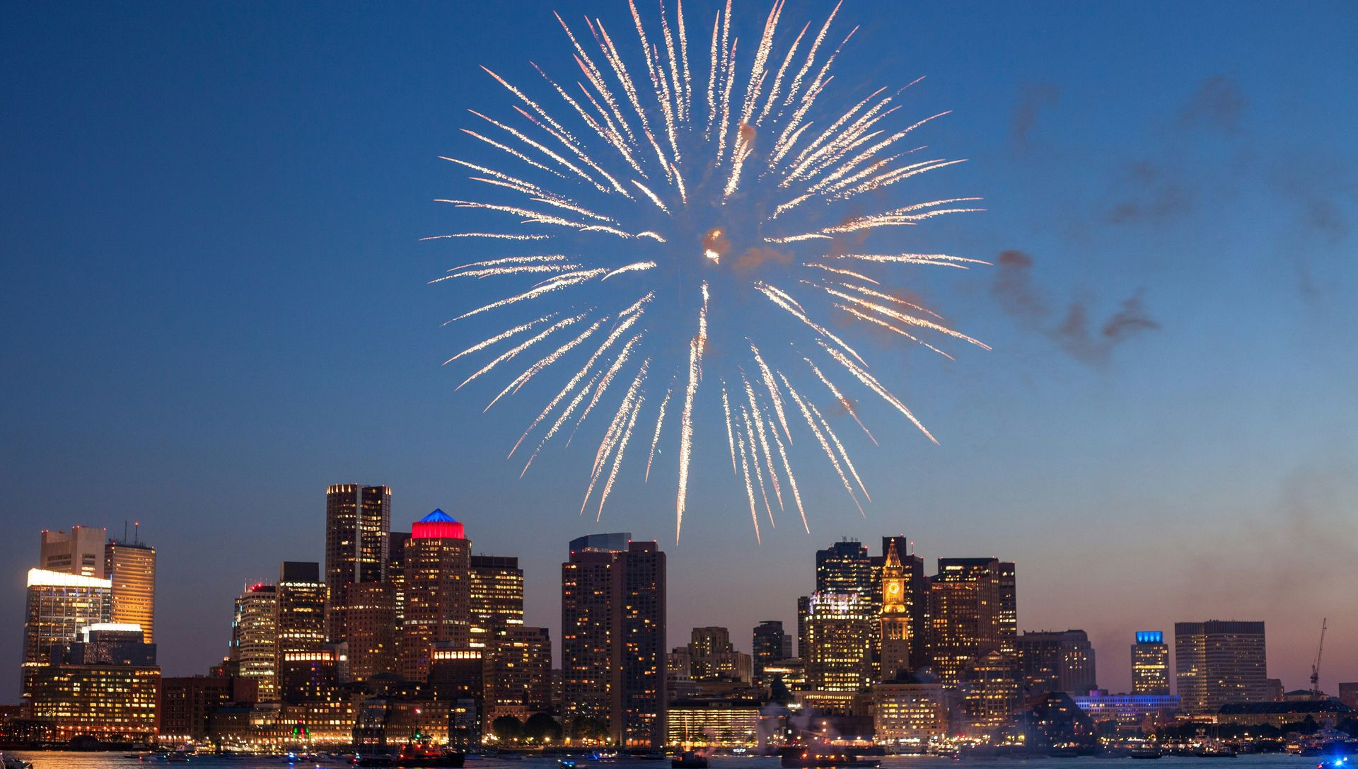 A brilliant white firework burst illuminates the Boston city skyline over the water at dusk.