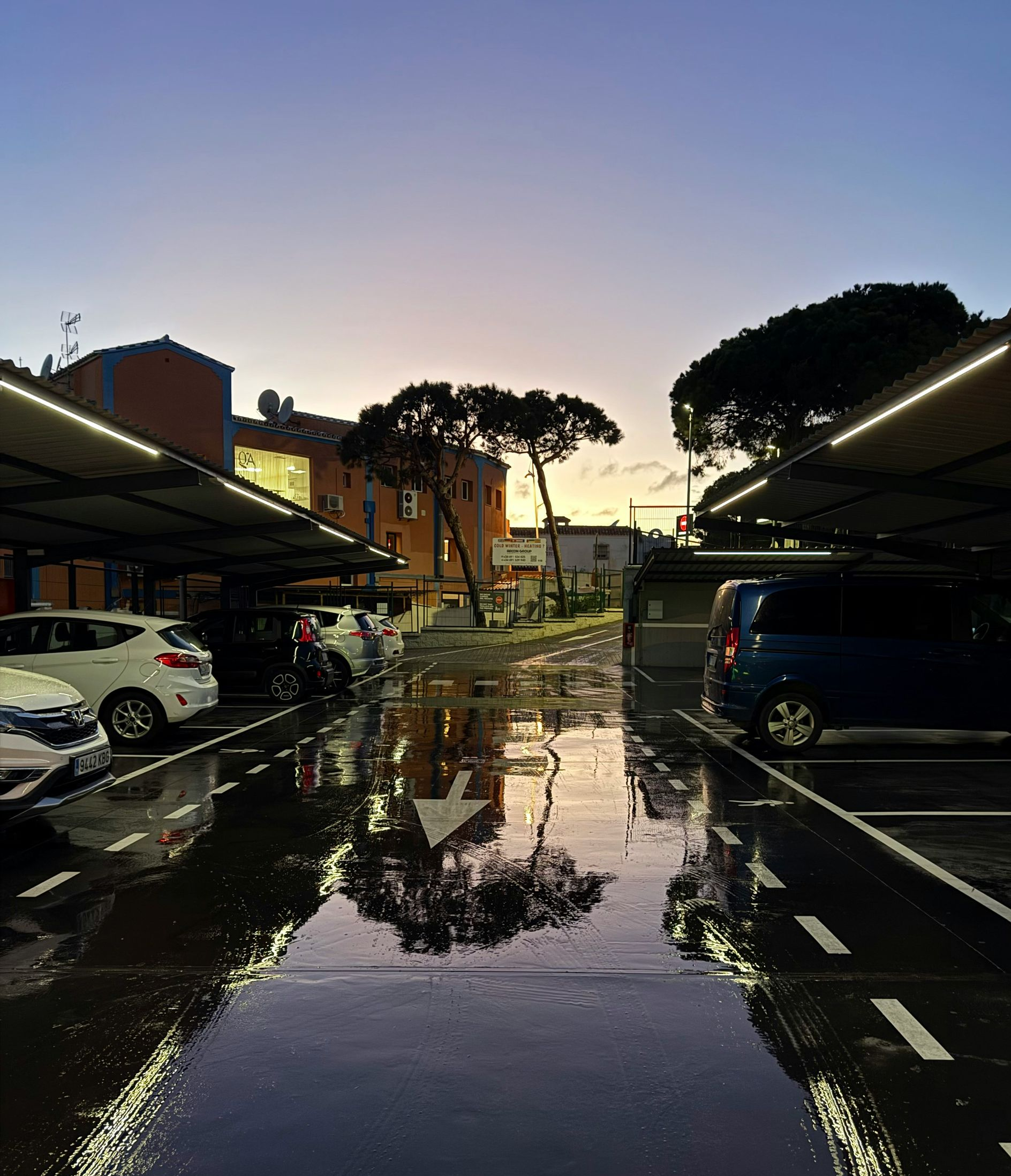 Rain-soaked parking lot at dusk, with wet pavement reflecting lights and parked cars