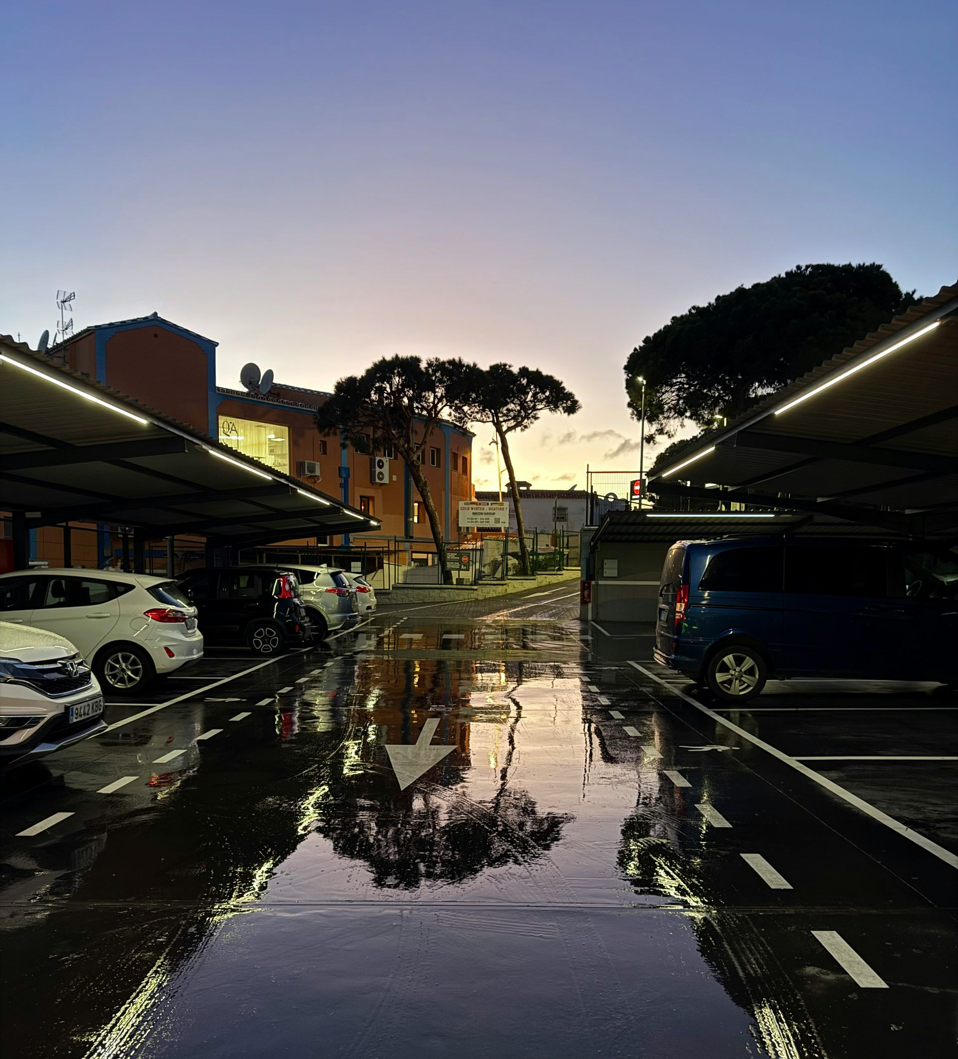 Rain-soaked parking lot at dusk, with wet pavement reflecting lights and parked cars