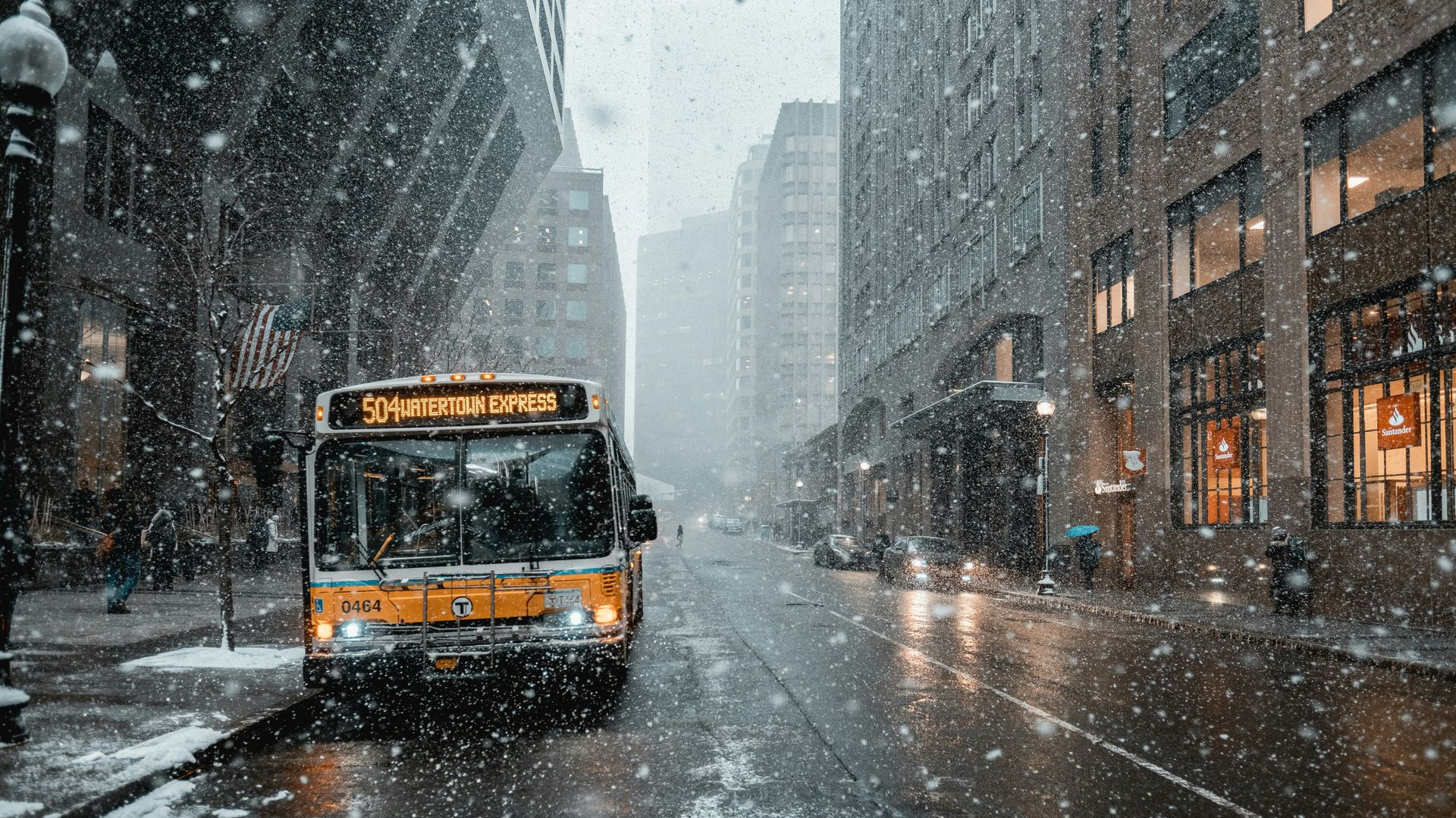 A yellow city bus travels down a snowy urban street lined with tall buildings.
