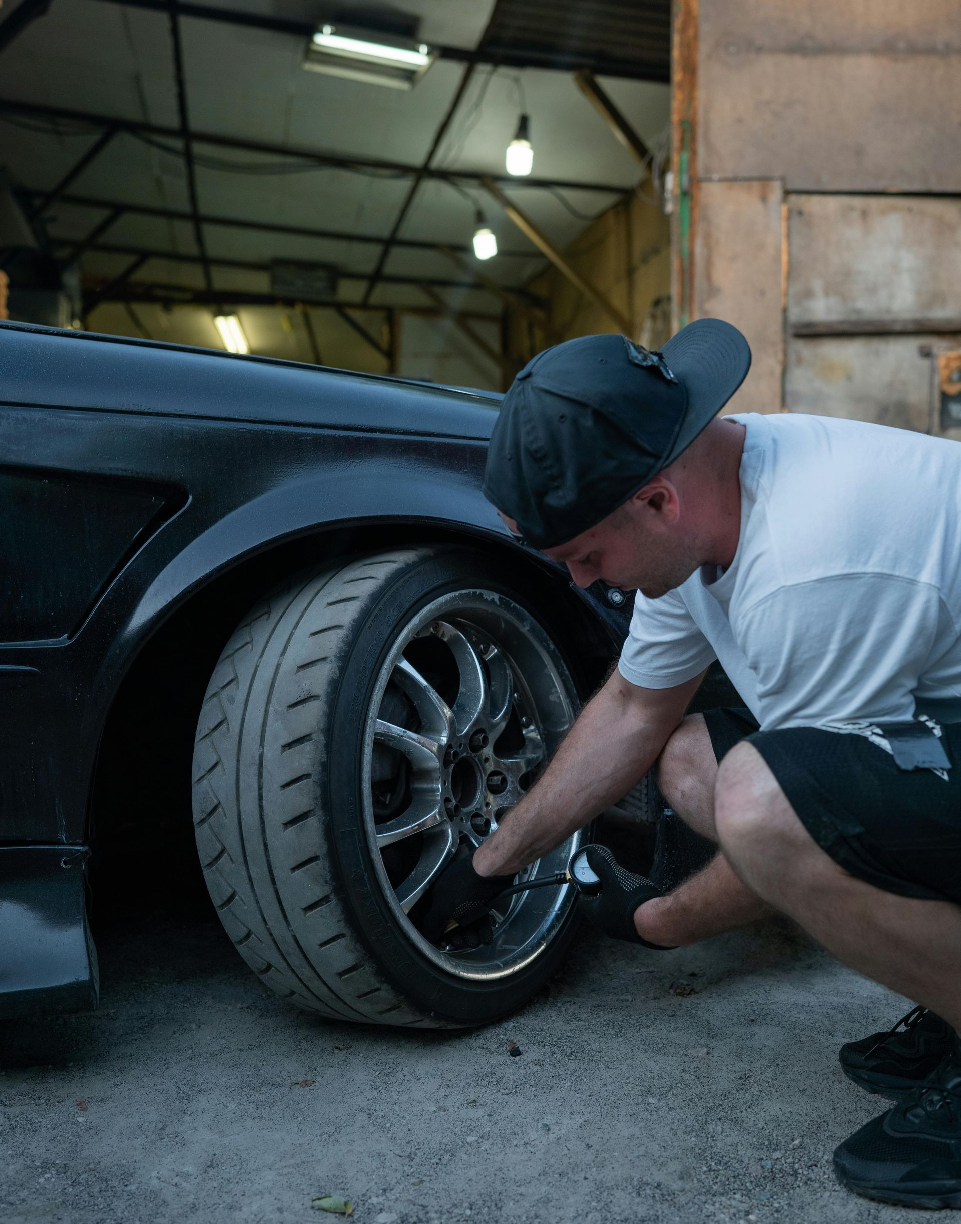 Man checking the wheel of a black car in a garage. He’s wearing a hat, shorts, and a white t-shirt.