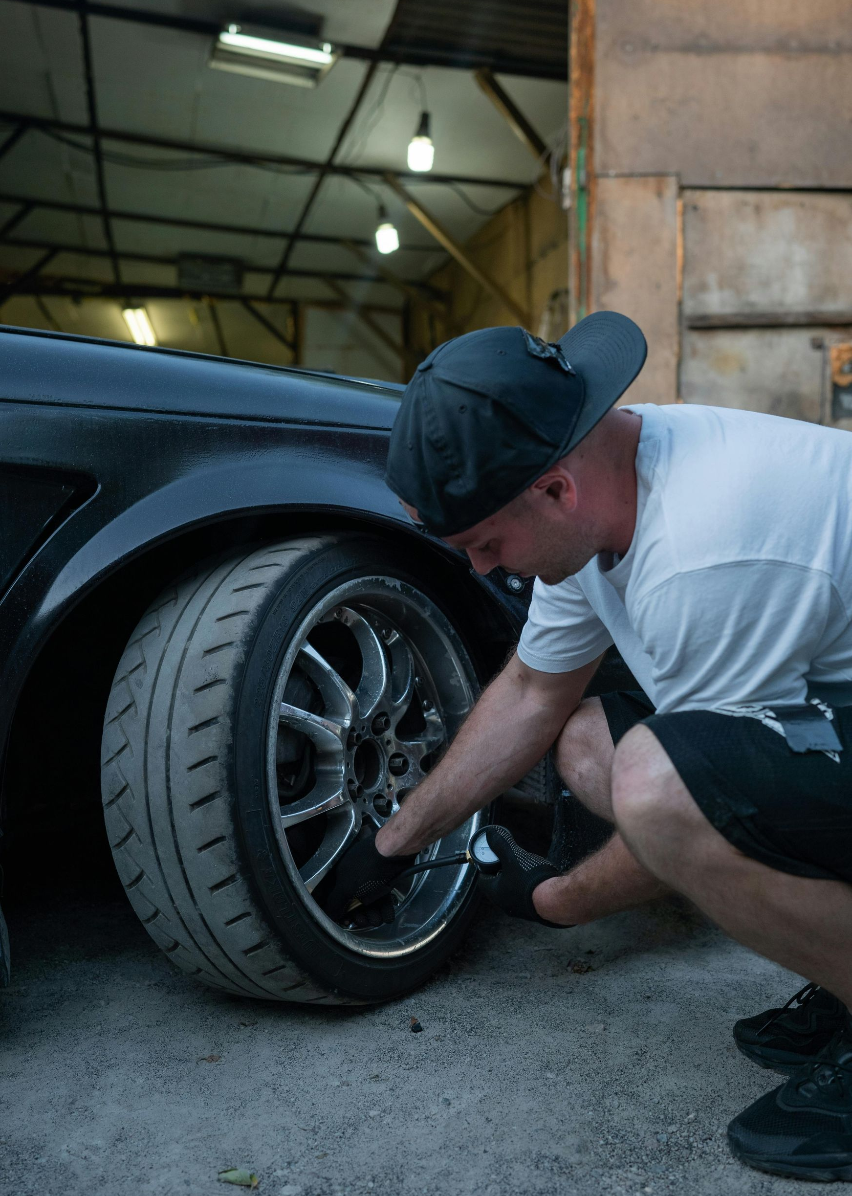 Man checking the wheel of a black car in a garage. He’s wearing a hat, shorts, and a white t-shirt.