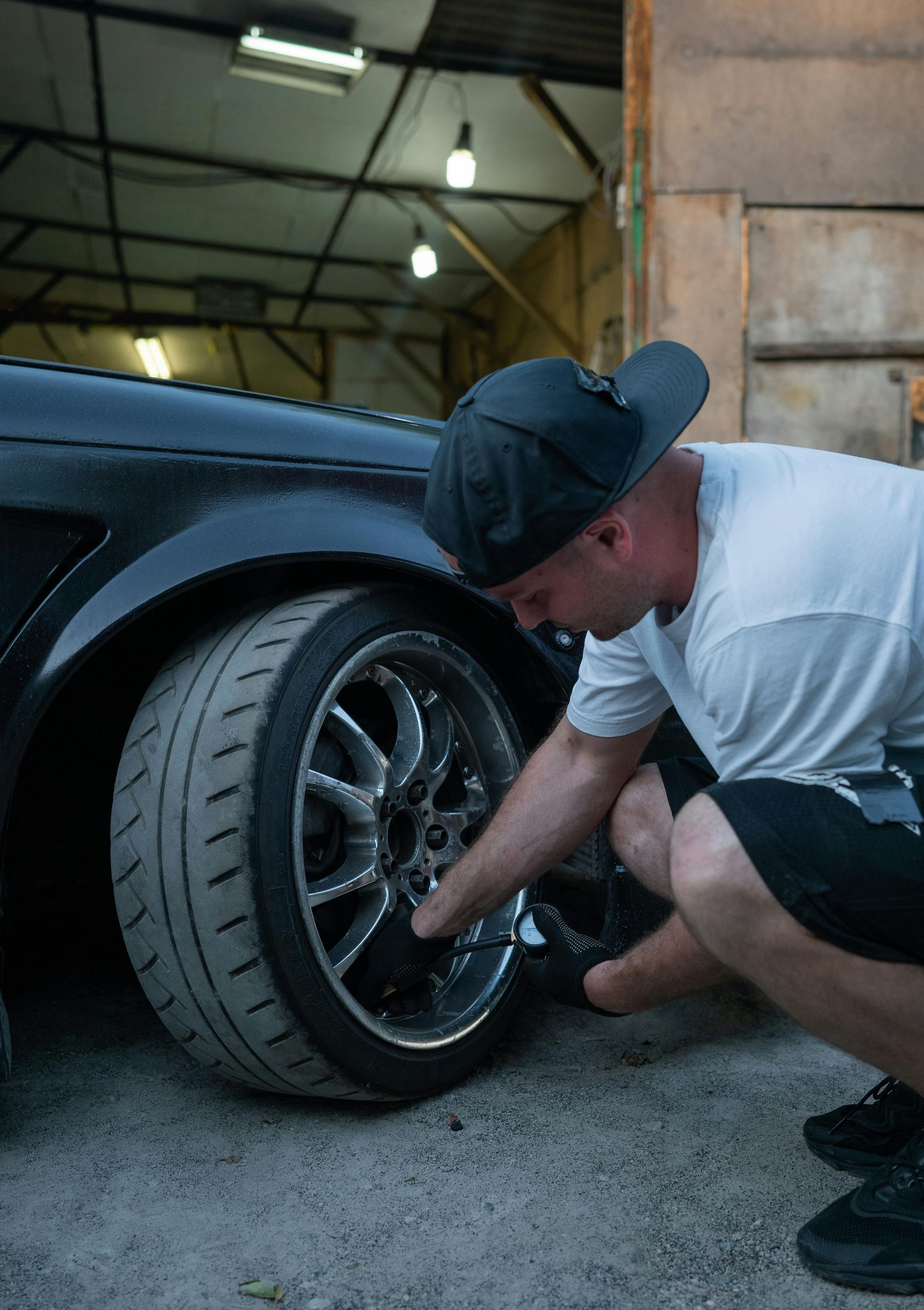Man checking the wheel of a black car in a garage. He’s wearing a hat, shorts, and a white t-shirt.