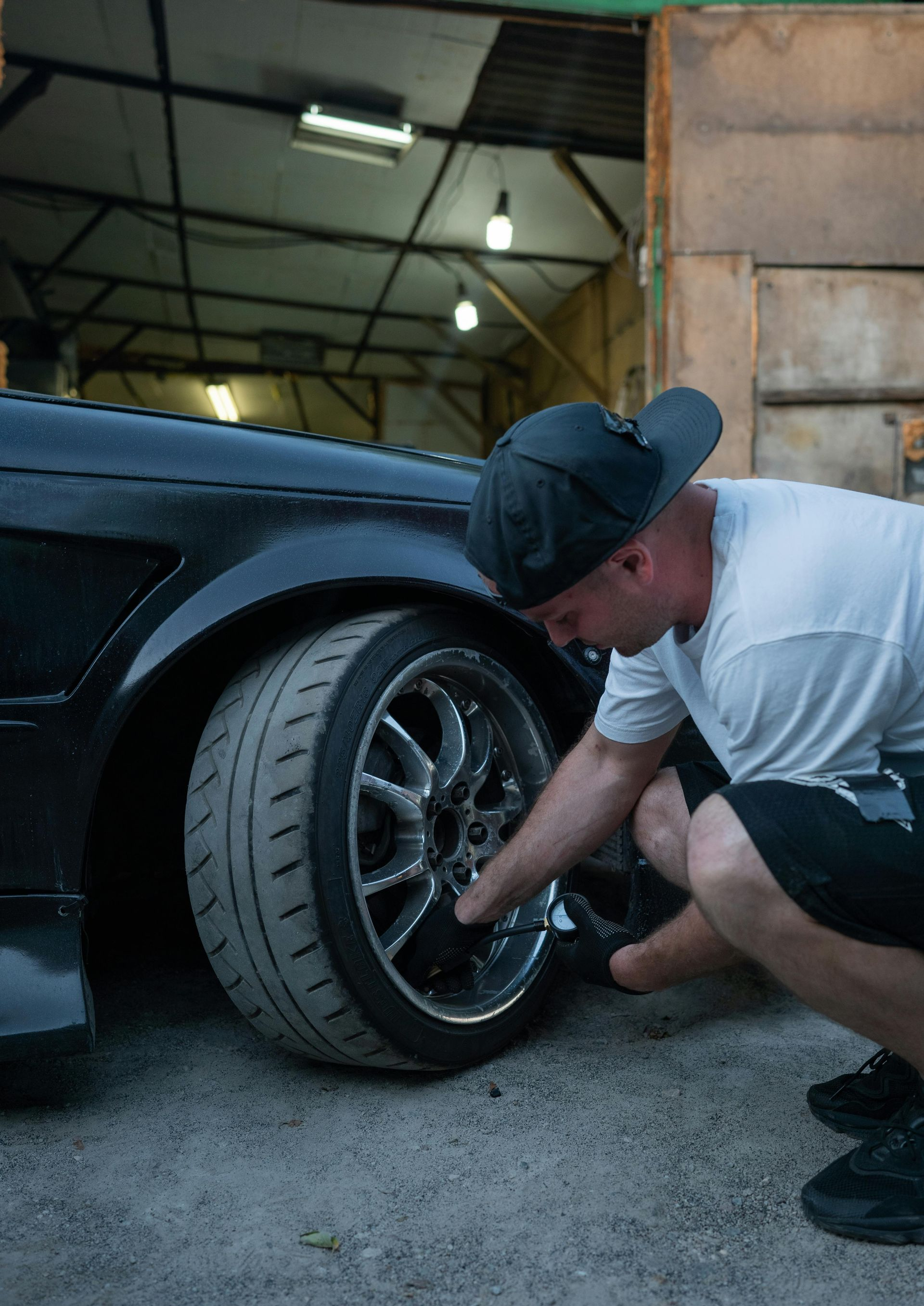 Man checking the wheel of a black car in a garage. He’s wearing a hat, shorts, and a white t-shirt.