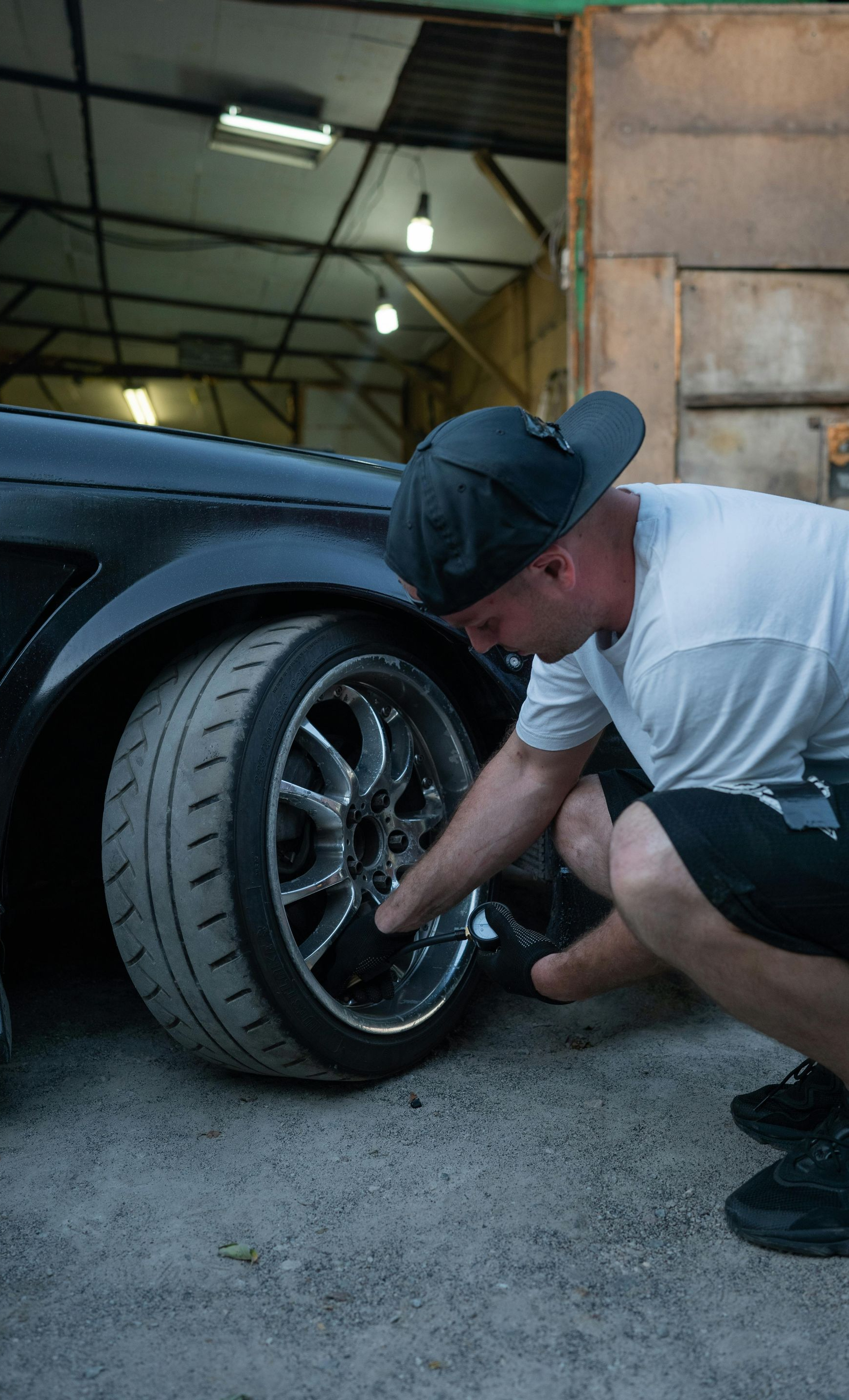 Man checking the wheel of a black car in a garage. He’s wearing a hat, shorts, and a white t-shirt.