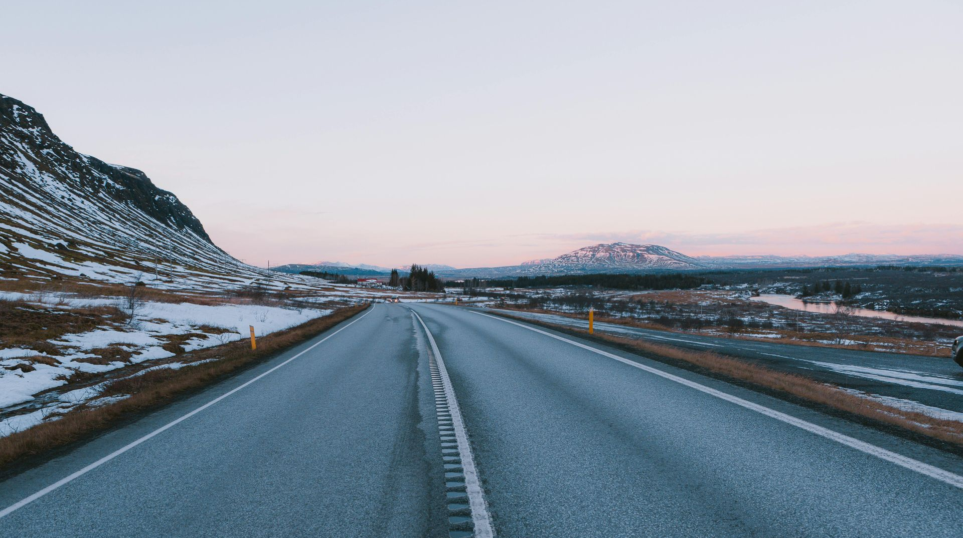 Empty highway through a mountainous, snow-covered landscape under a pale sunset sky.