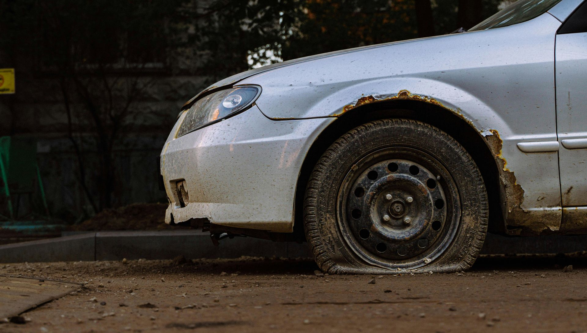 A close-up of a white car with a flat tire on a dirt road, showing rust on the fender.