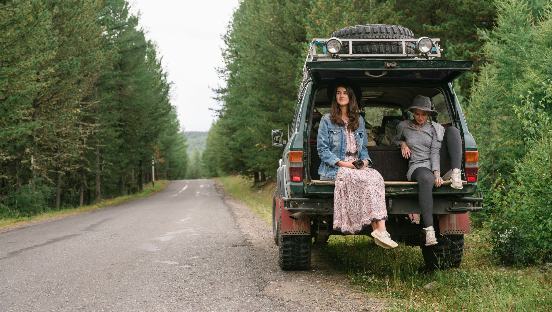 Two people sitting on the back of an SUV on a forest road, with one person smiling.