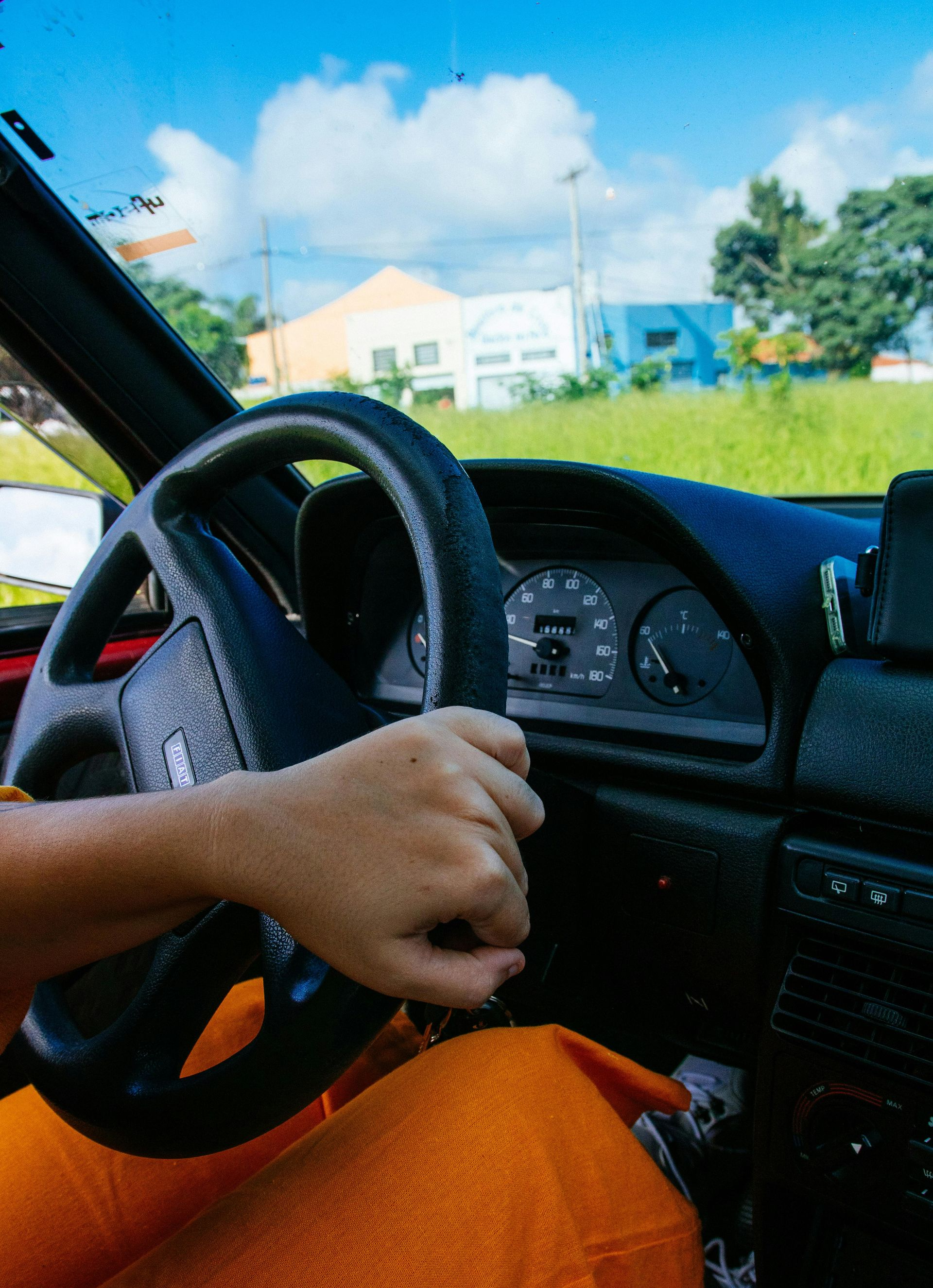 Person's hand on a car steering wheel, driving on a sunny day. Interior view, orange pants visible.