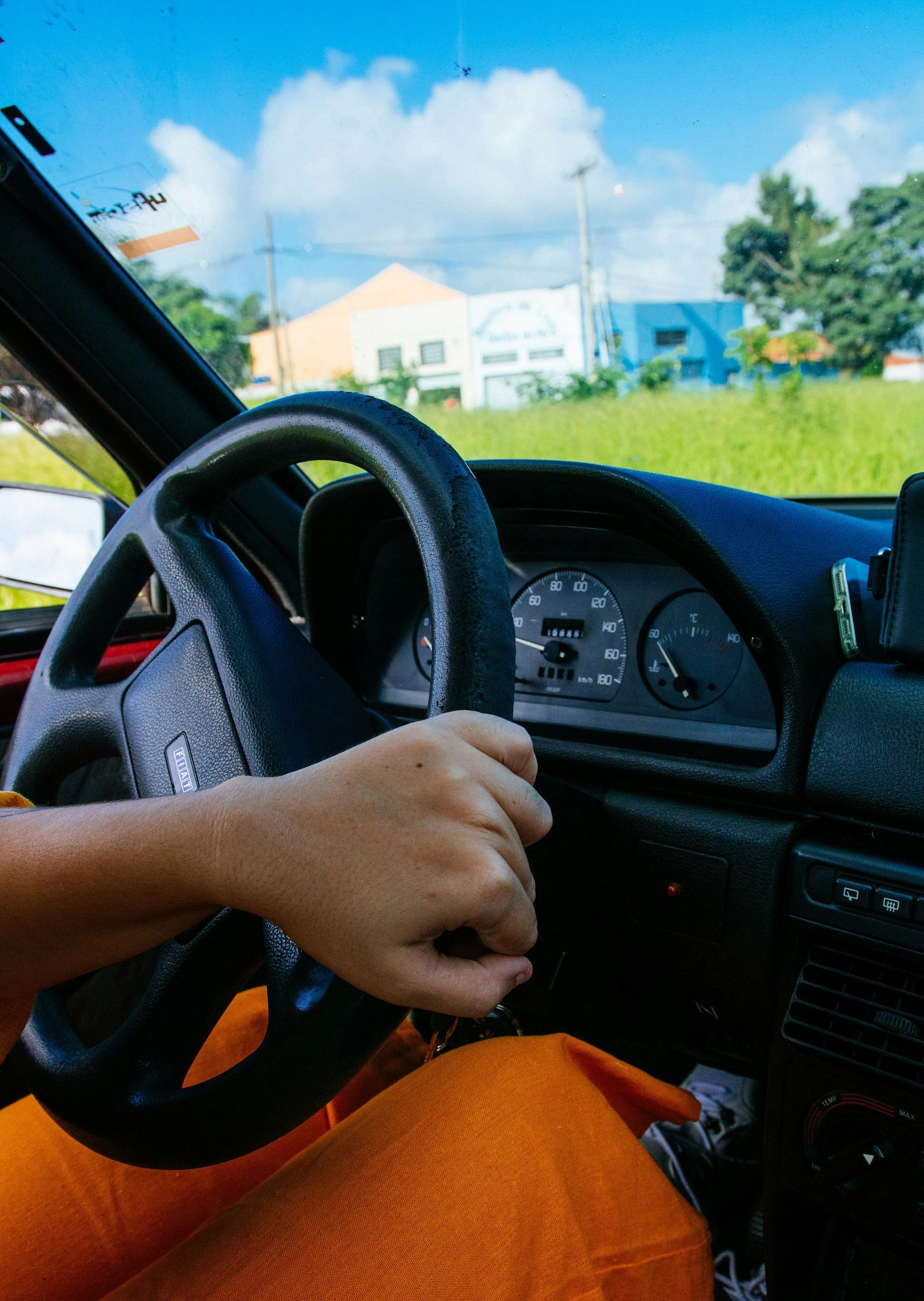 Person's hand on a car steering wheel, driving on a sunny day. Interior view, orange pants visible.