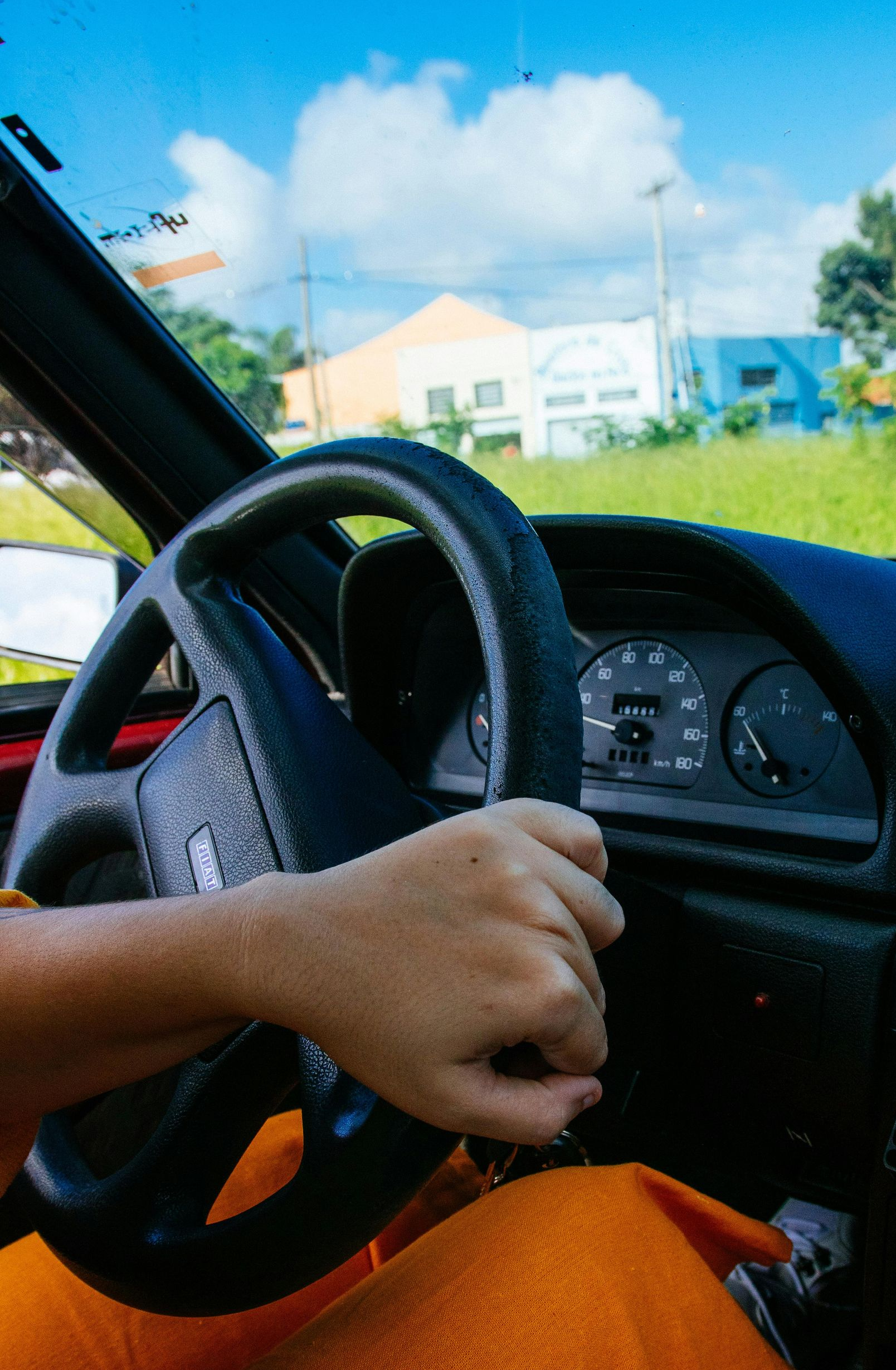 Person's hand on a car steering wheel, driving on a sunny day. Interior view, orange pants visible.