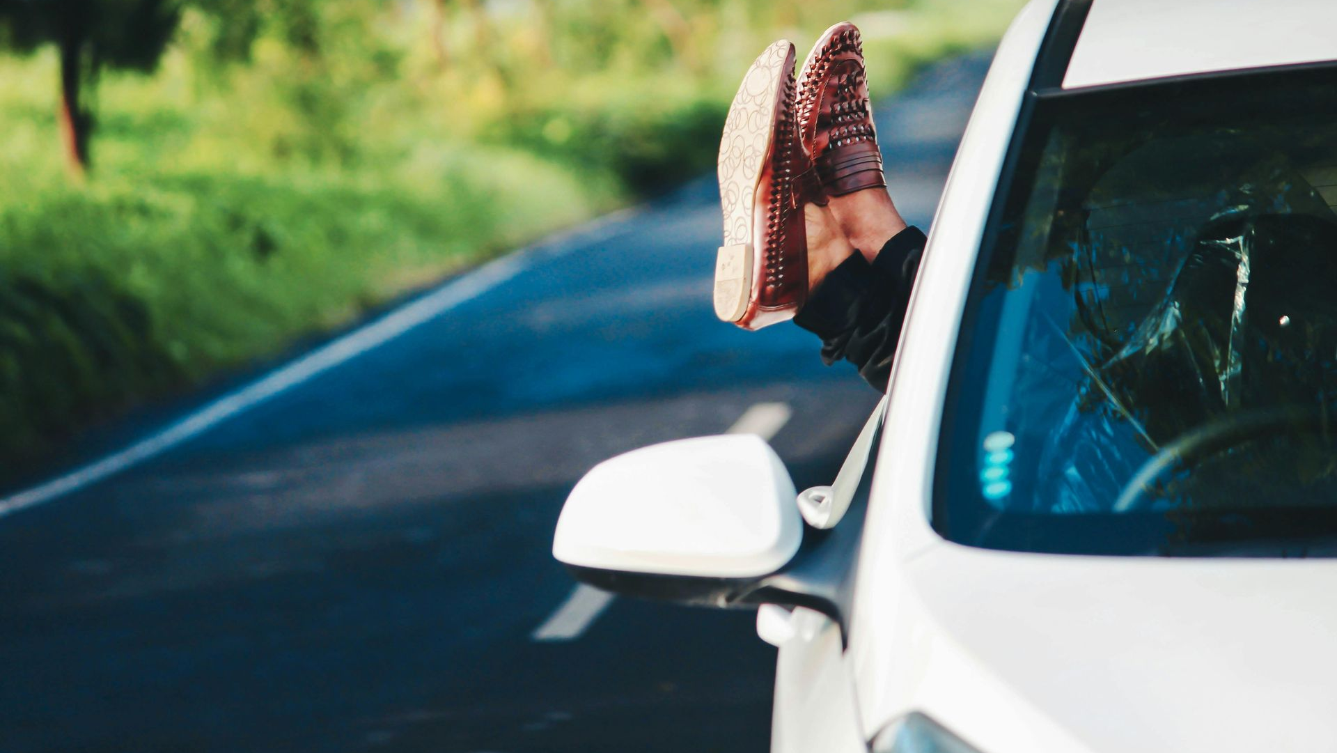 Person relaxing in a white car, feet out the window. Driving on a road with green foliage.