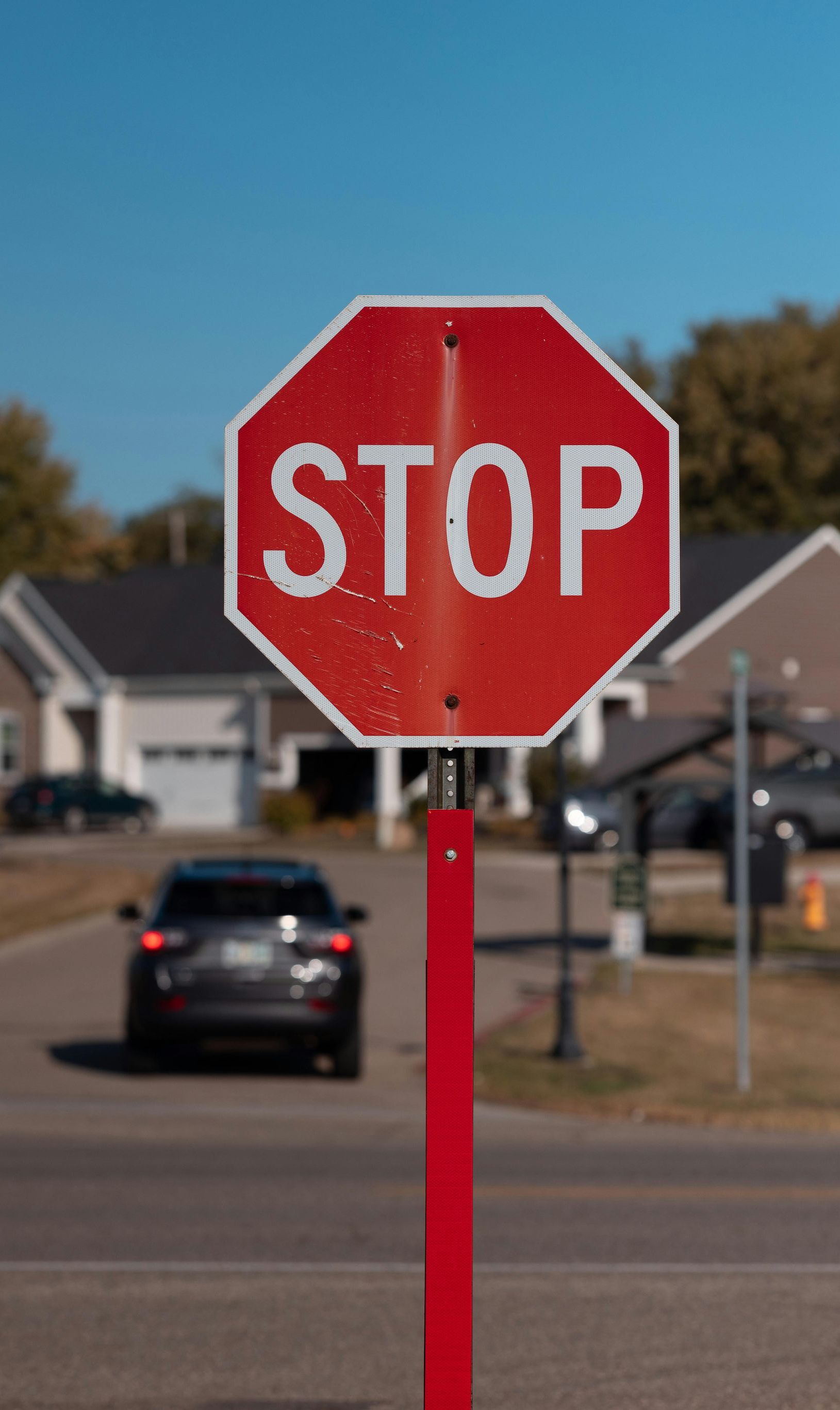 Red stop sign on a red pole in front of a street with a car and houses on a sunny day.