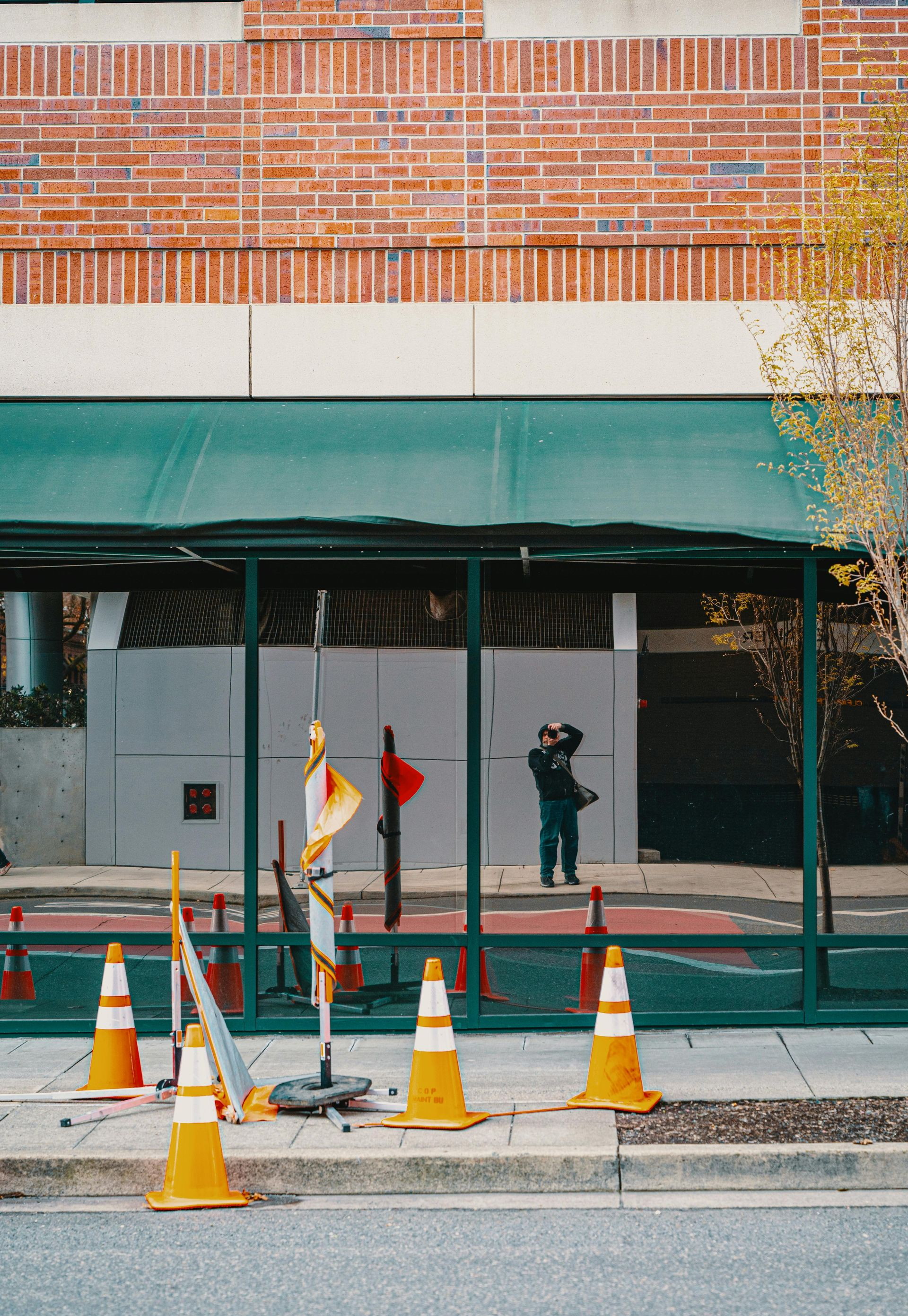 Construction cones and barriers in front of a building with green awning and reflective glass.
