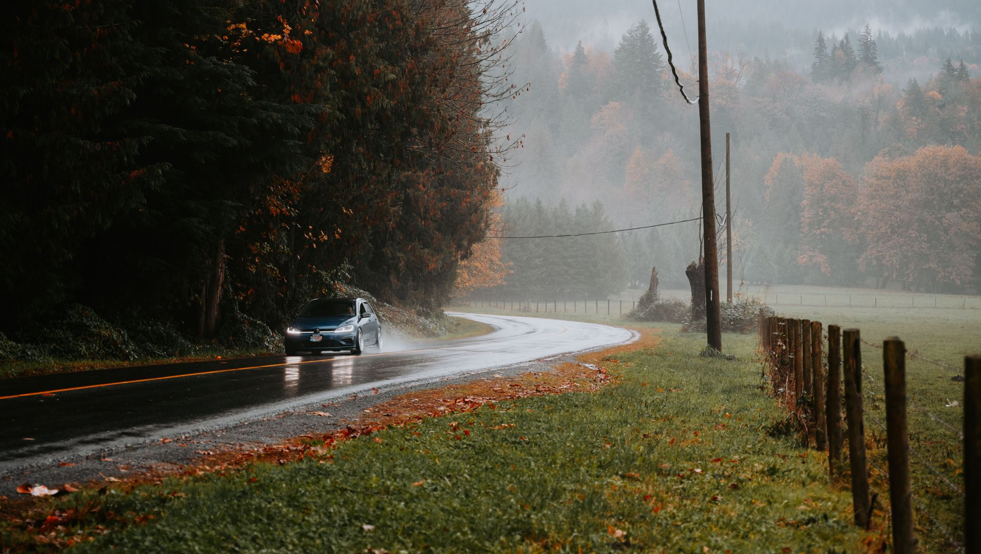 Rainy rural road with a car driving through mist beside autumn trees and a fenced field